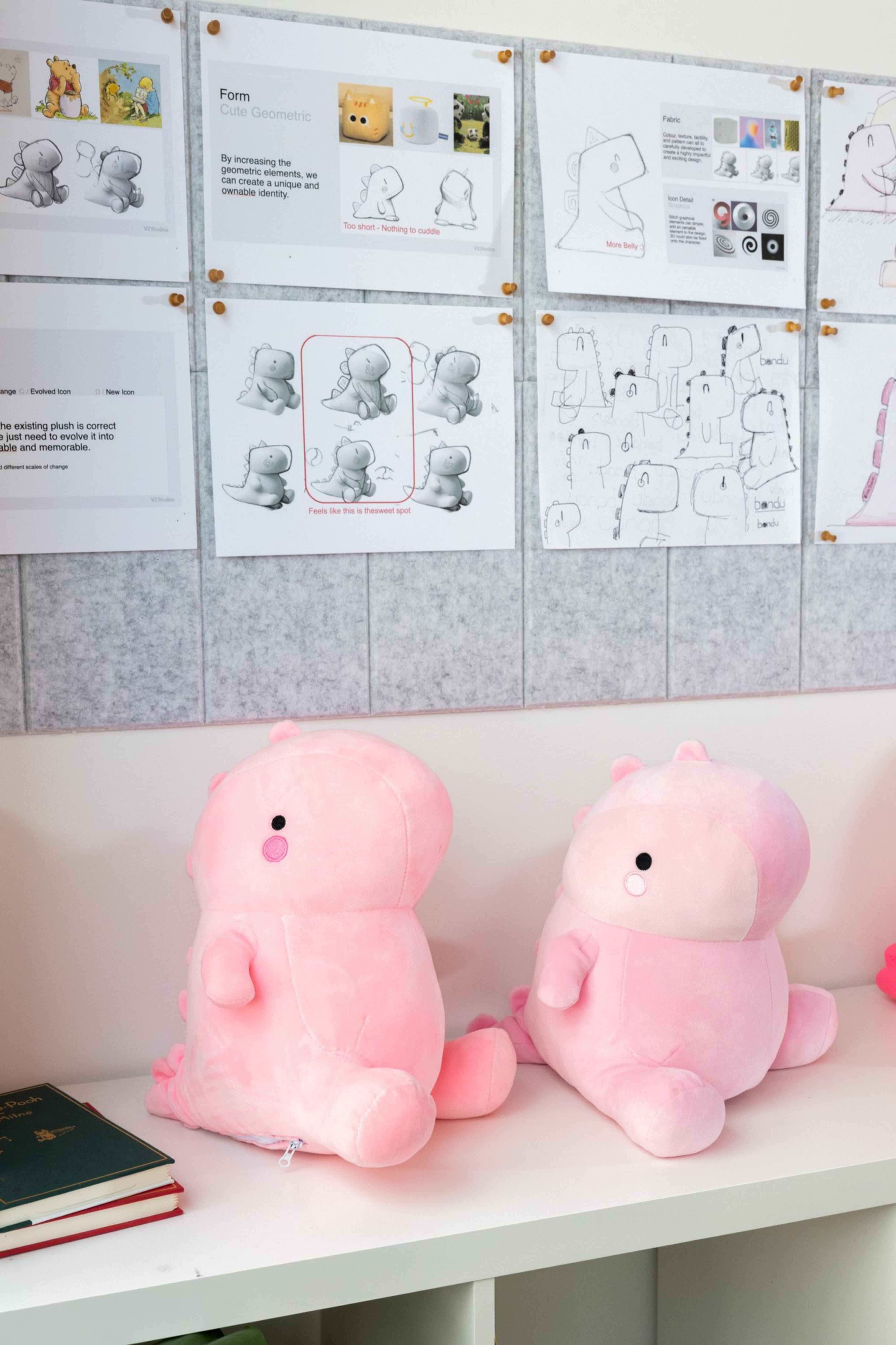 Two pink dinosaur plush toys sit side by side on a white shelf below a board with sketches, diagrams, and instructions for designing the plush.