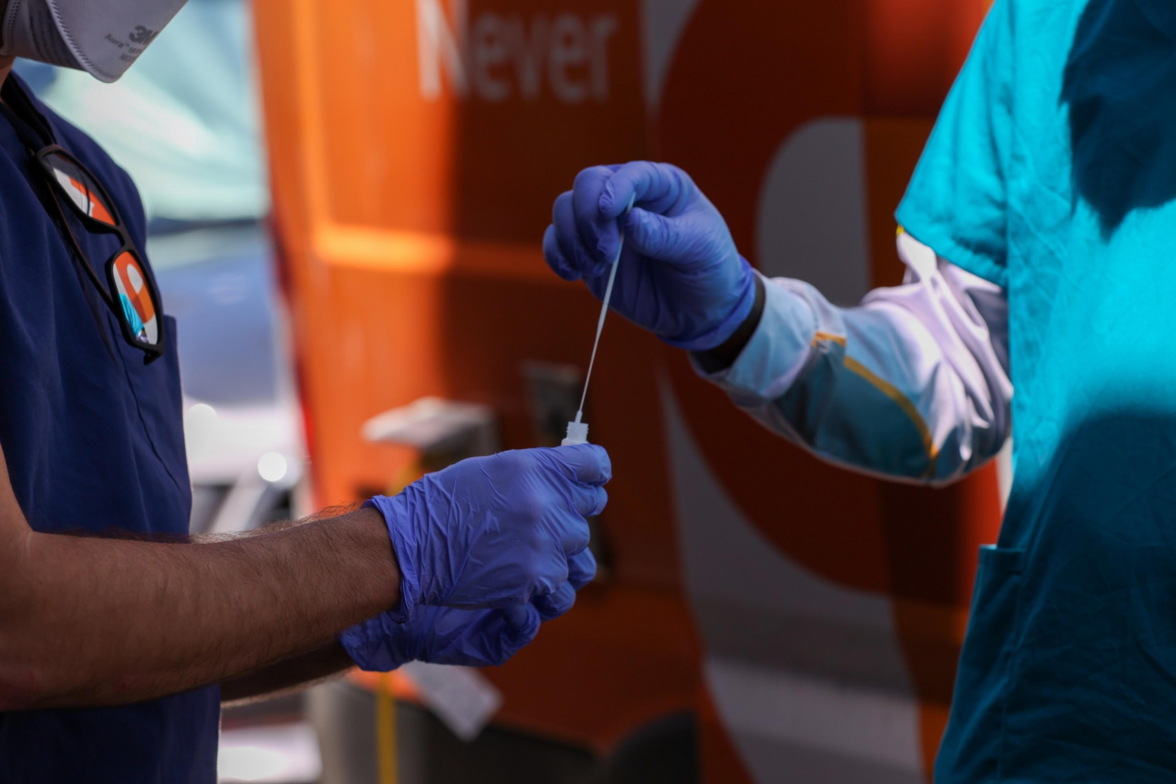 Two people wearing blue gloves exchange a COVID-19 test swab outdoors near an orange vehicle.