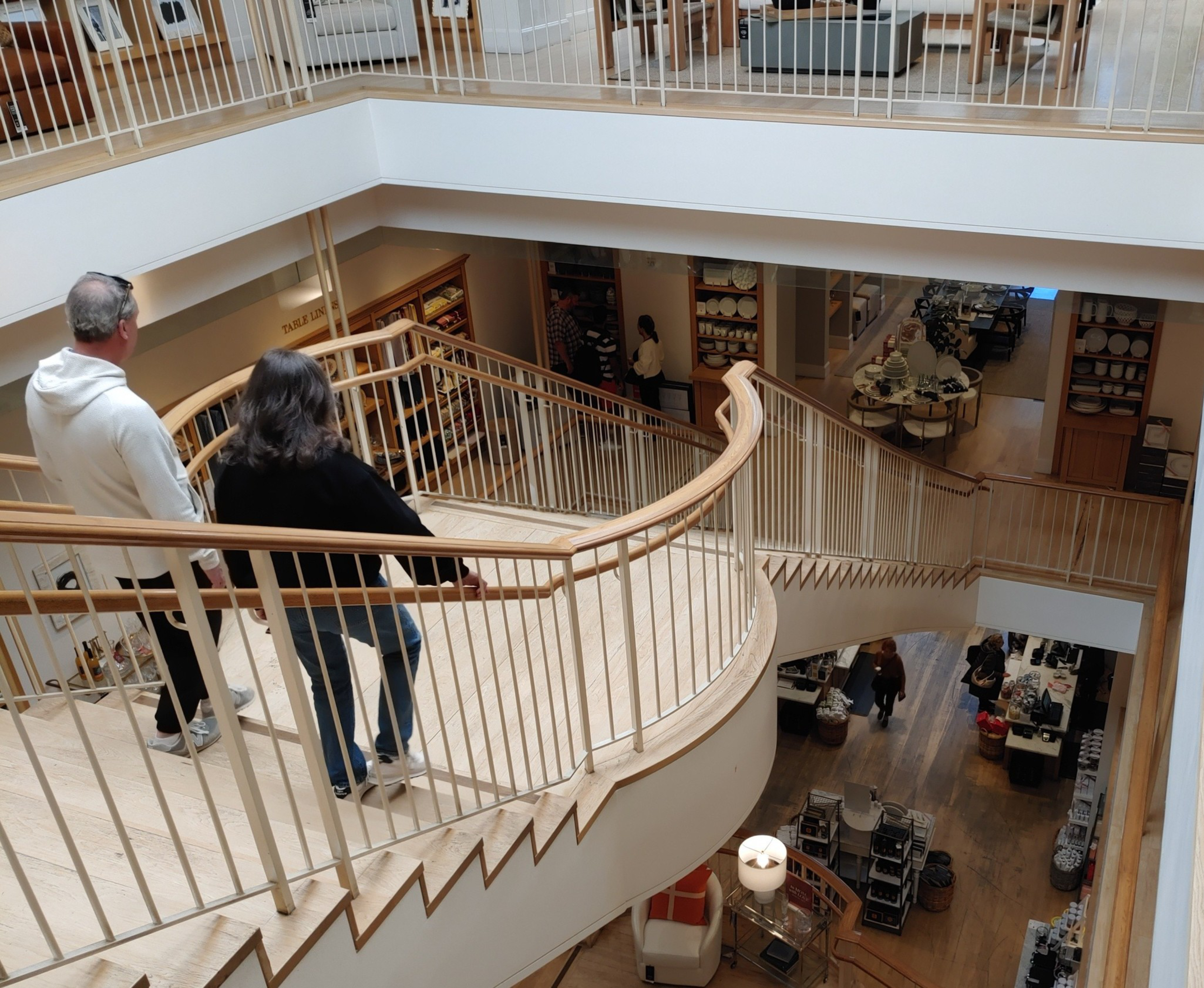 A man and a woman walk down a wide wooden staircase with metal railings inside a multi-story store filled with shelves and display tables.