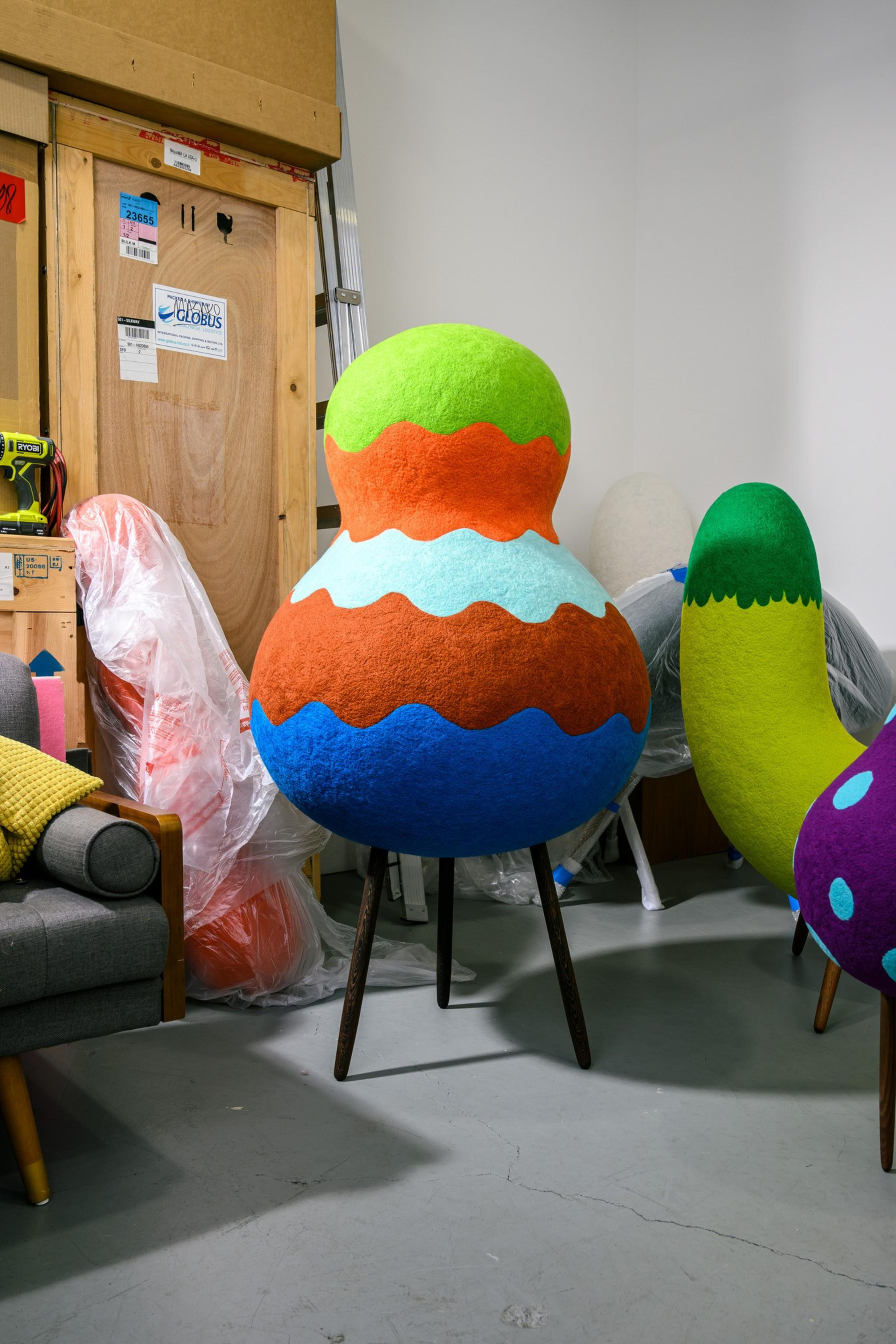The image shows colorful, abstract sculptures with wavy patterns on legs in a cluttered room with boxes and a gray chair nearby.