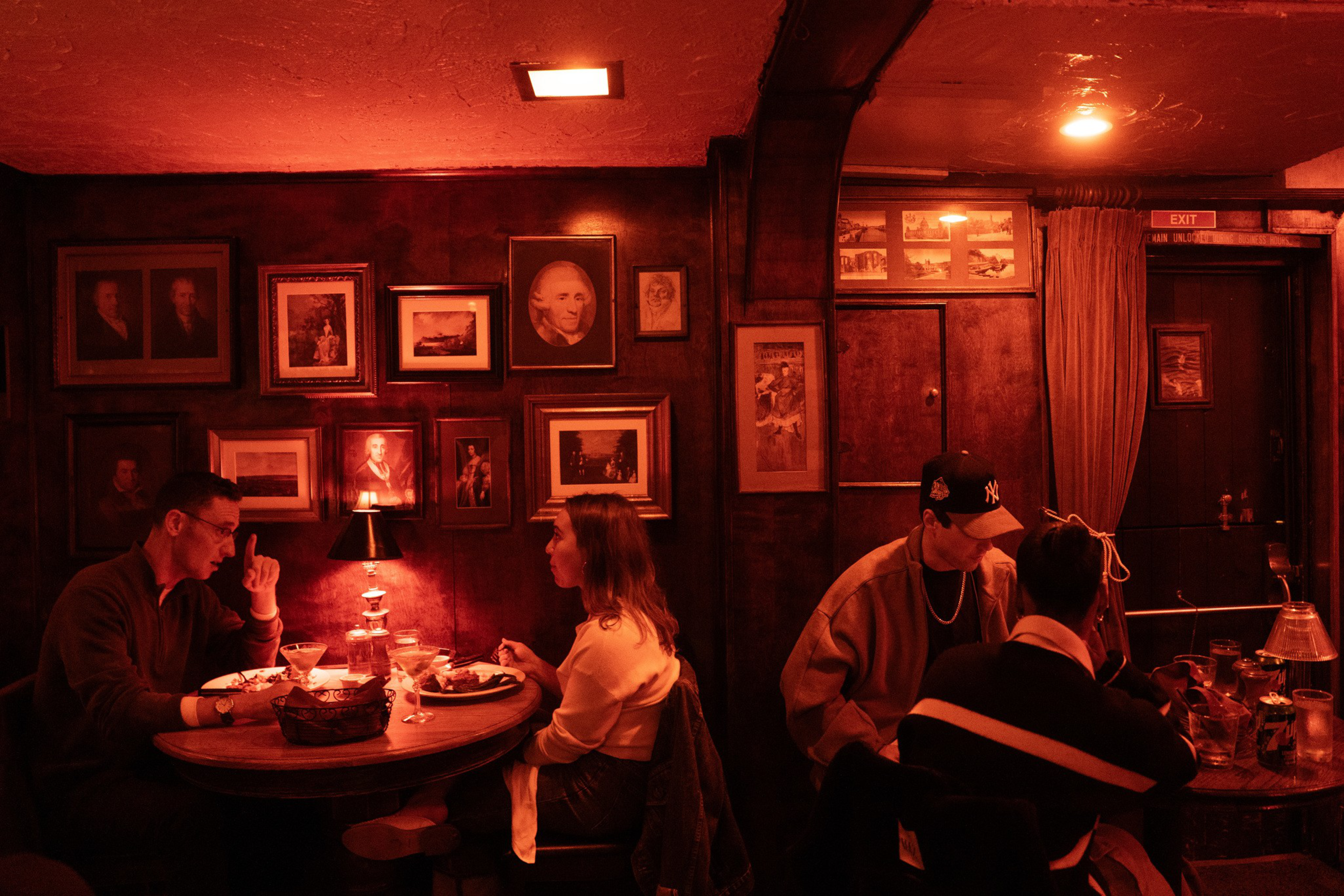 Two couples sit at separate wooden tables in a dimly lit bar with red lighting and walls adorned with framed vintage portraits and paintings.