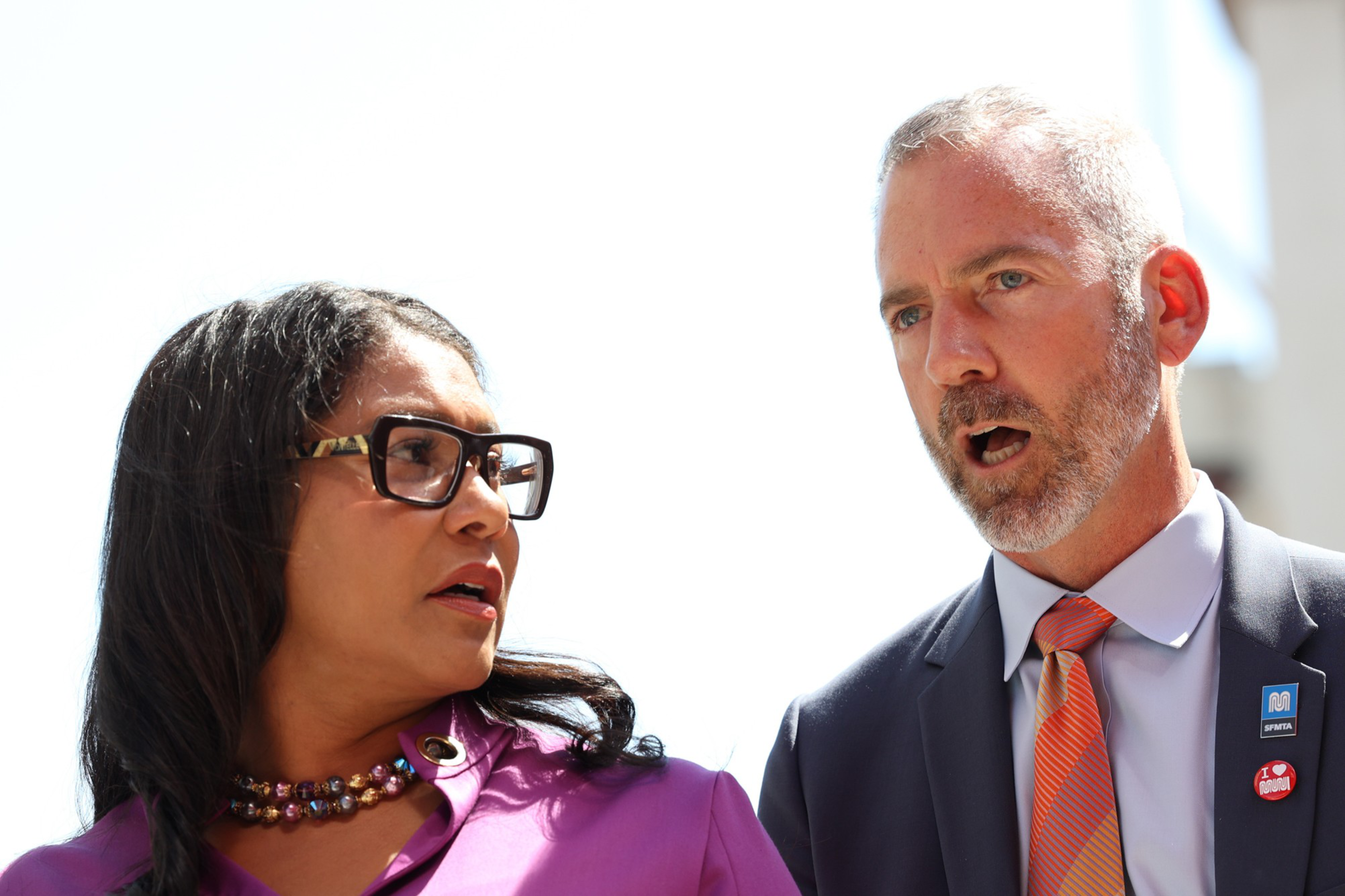 Mayor London Breed and SFMTA Director of Transportation Jeffrey Tumlin are engaged in conversation. The woman, with long black hair, wears glasses, a purple shirt, and a colorful necklace. The man, with gray hair and beard, is in a suit with pins.