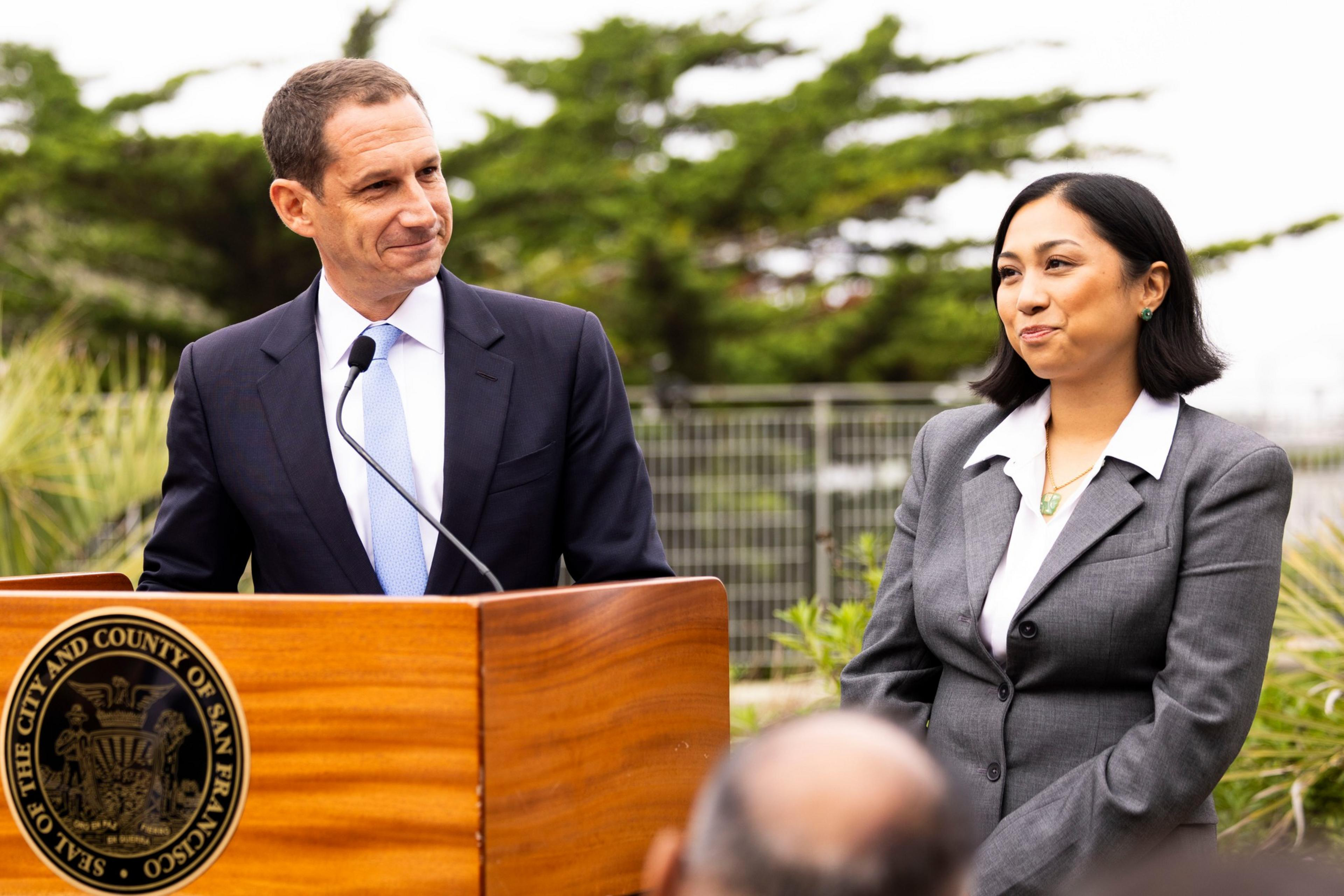 A man in a dark suit and blue tie speaks at a wooden podium while a woman in a gray suit stands beside him, both smiling outdoors.