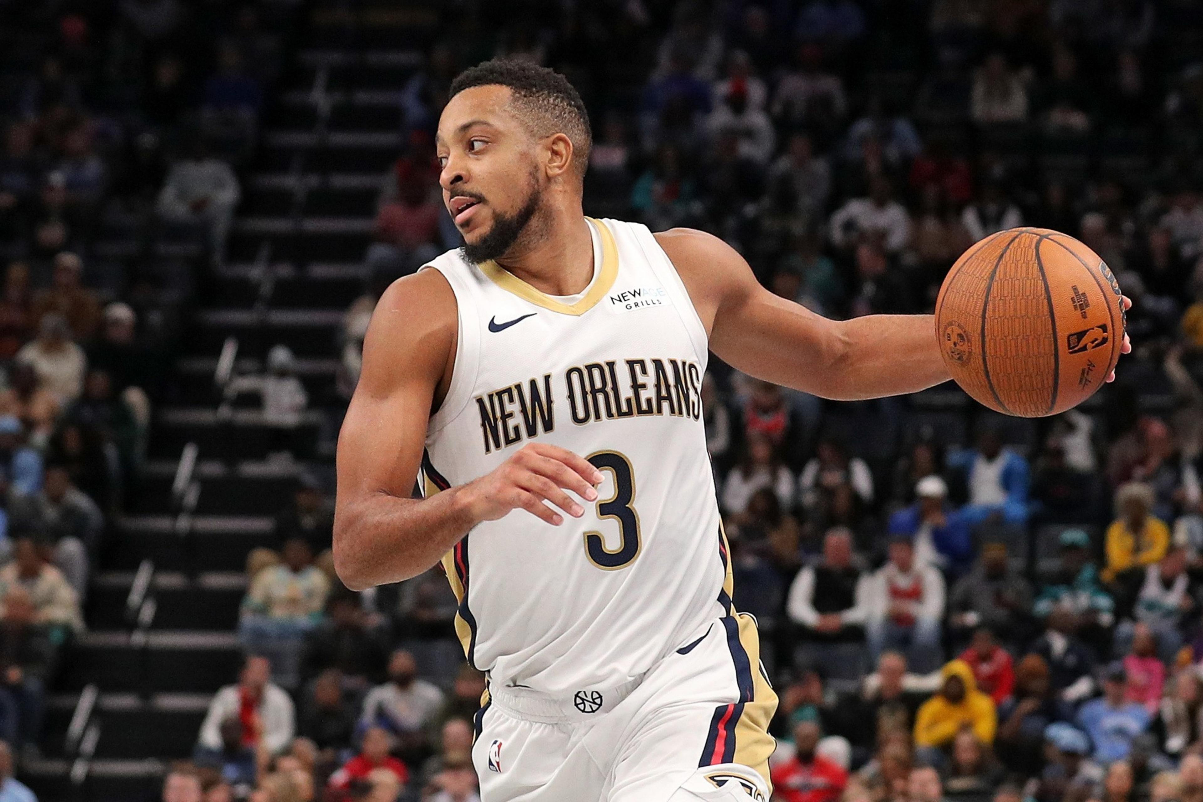 A basketball player in a New Orleans jersey is dribbling a ball on a court with a focused expression. The crowd watches in the background.