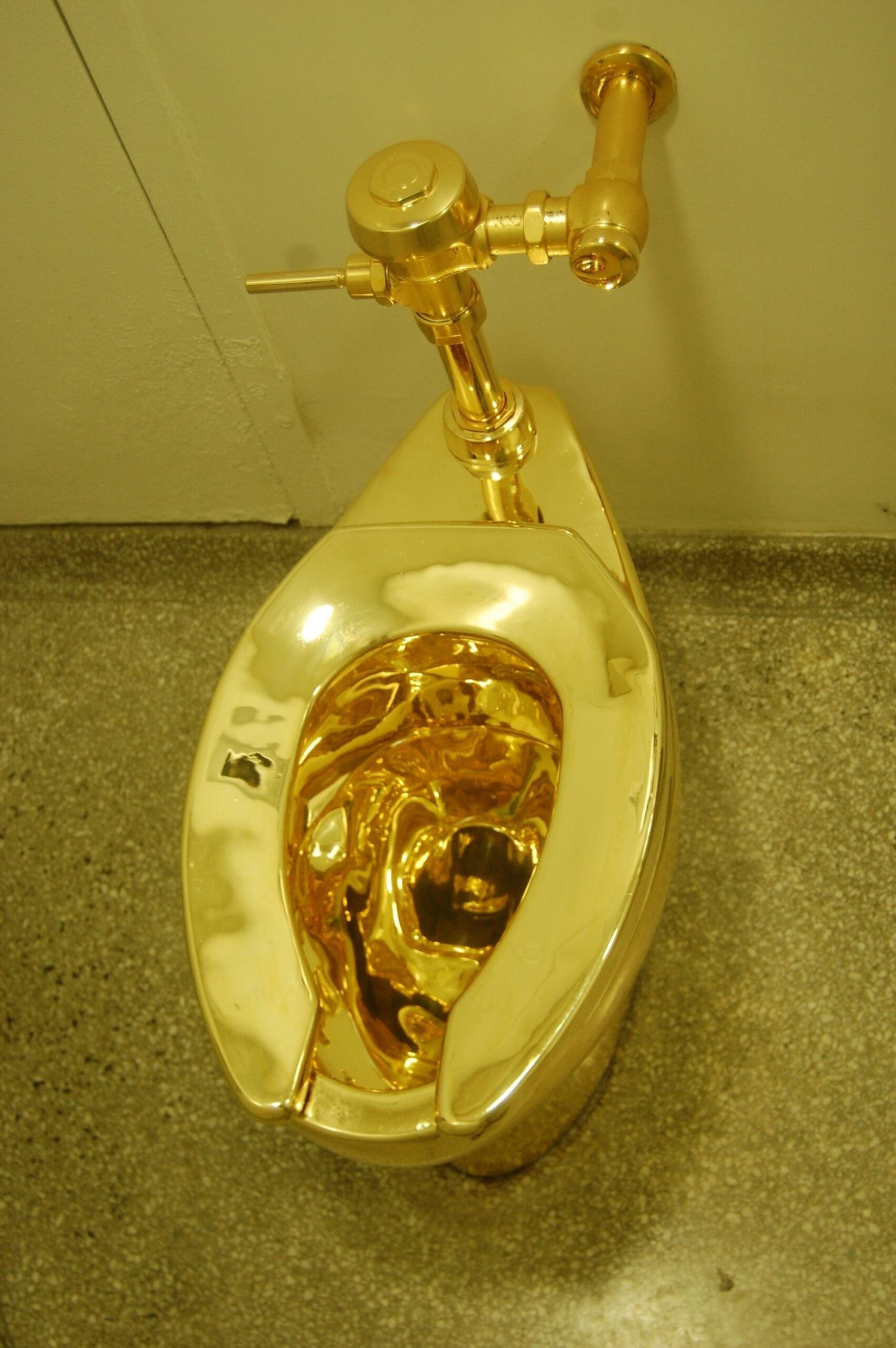 A shiny gold-plated toilet with a matching gold flush valve mounted on a speckled floor against a plain wall.