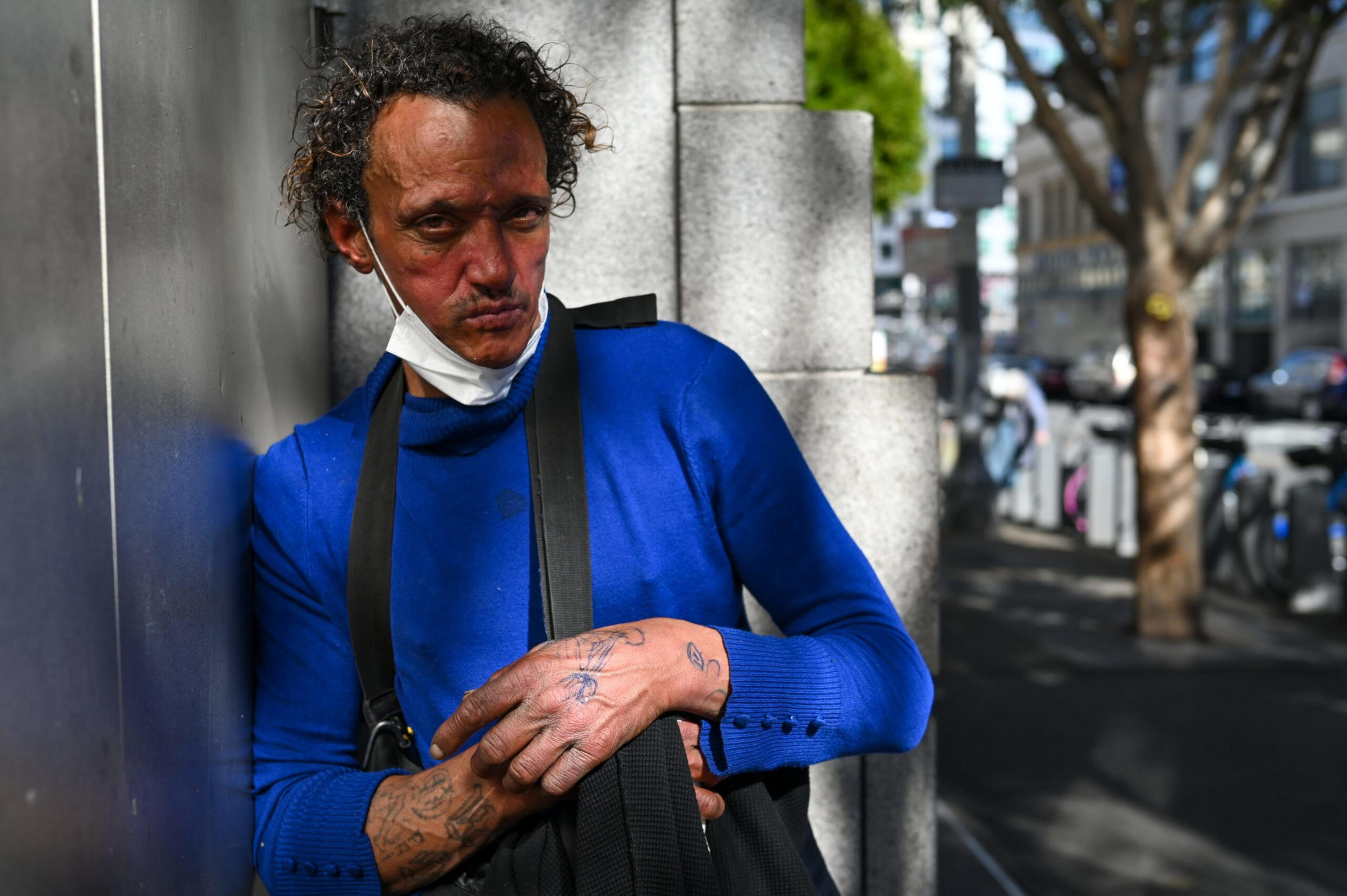 A man with curly hair leans against a wall wearing a bright blue sweater and a partially lowered white face mask, with tattoos visible on his hands.