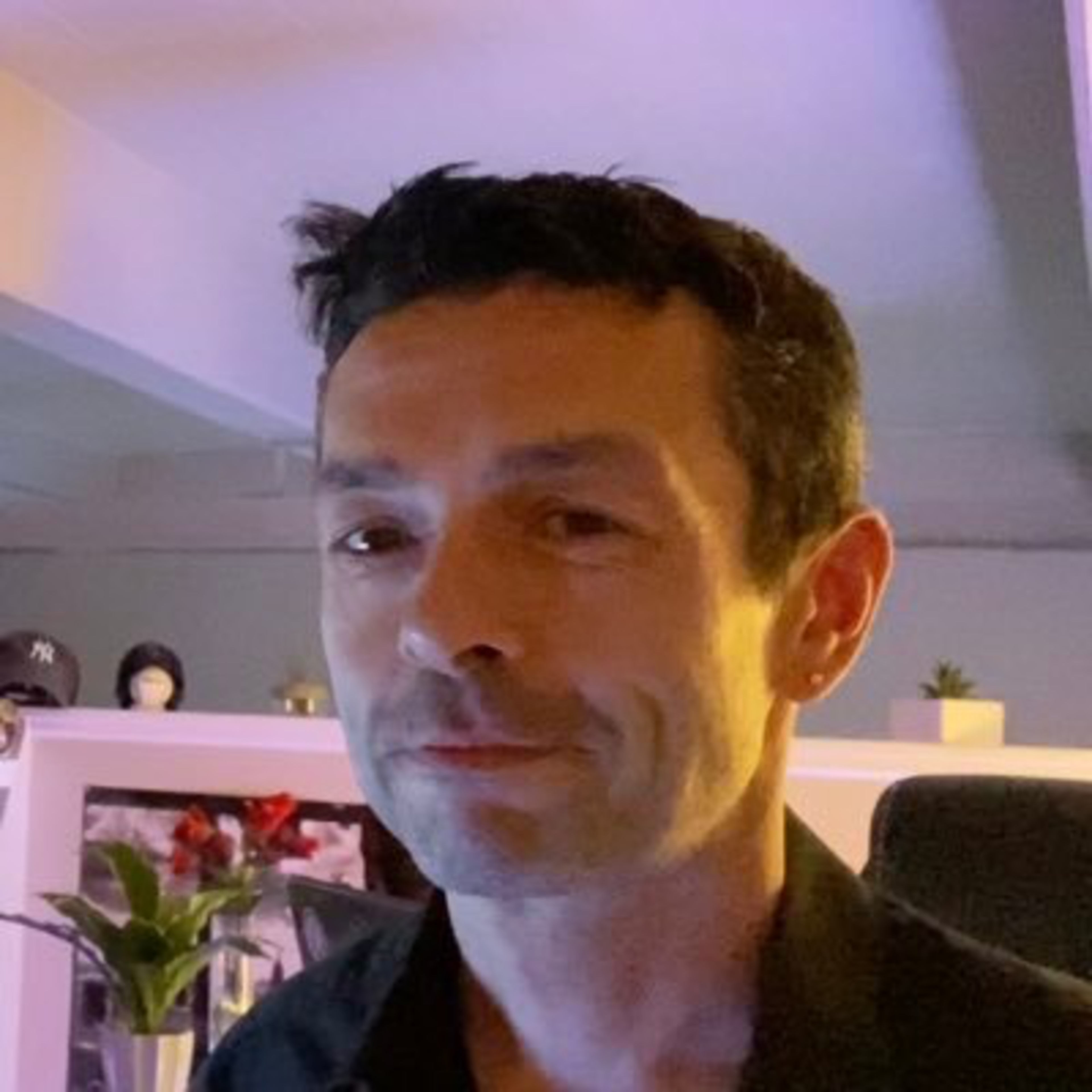 A man with short dark hair and a slight smile is indoors, wearing a black shirt, with plants and hats on shelves in a softly lit room behind him.