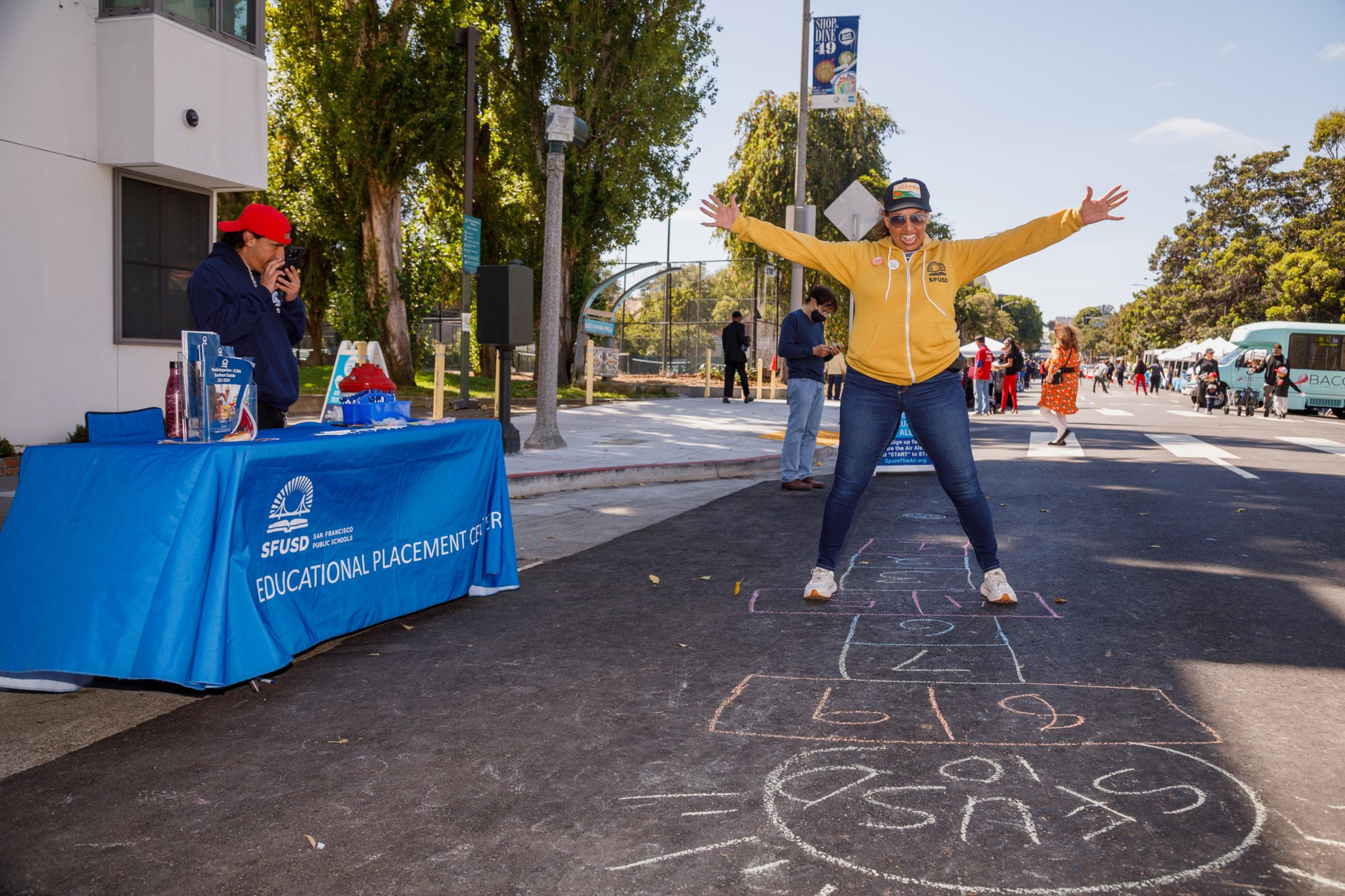A woman jumps on a hopscotch grid with arms wide open while a man stands behind a booth marked &quot;SFUSD.&quot; Trees line the sunny street.