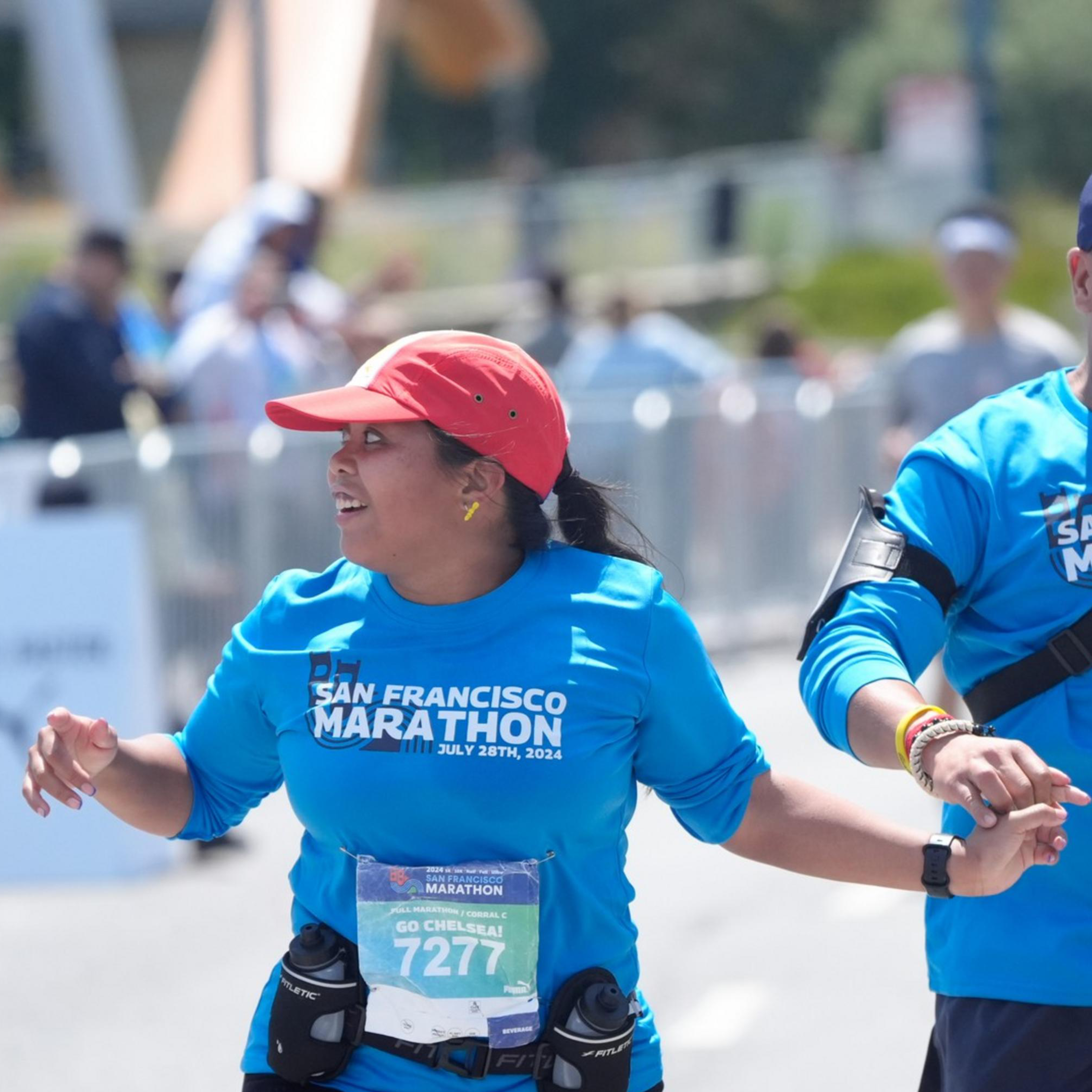 Two marathon runners, wearing blue &quot;San Francisco Marathon&quot; shirts, joyfully hold hands while running, surrounded by spectators and fellow participants.