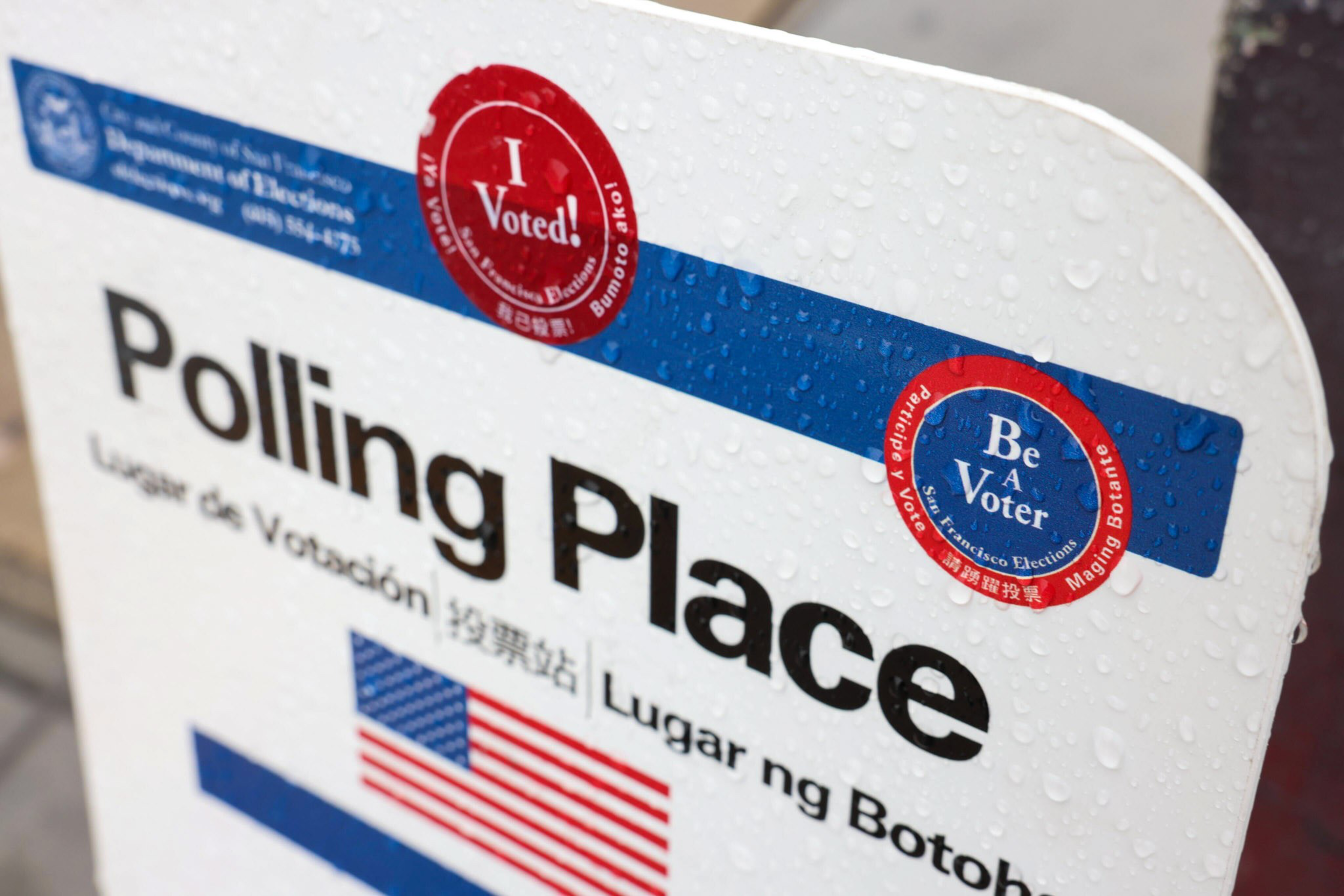 A rain-speckled "Polling Place" sign with "I Voted!" and "Be A Voter" stickers, featuring English, Spanish, Chinese, and Filipino text and a U.S. flag.