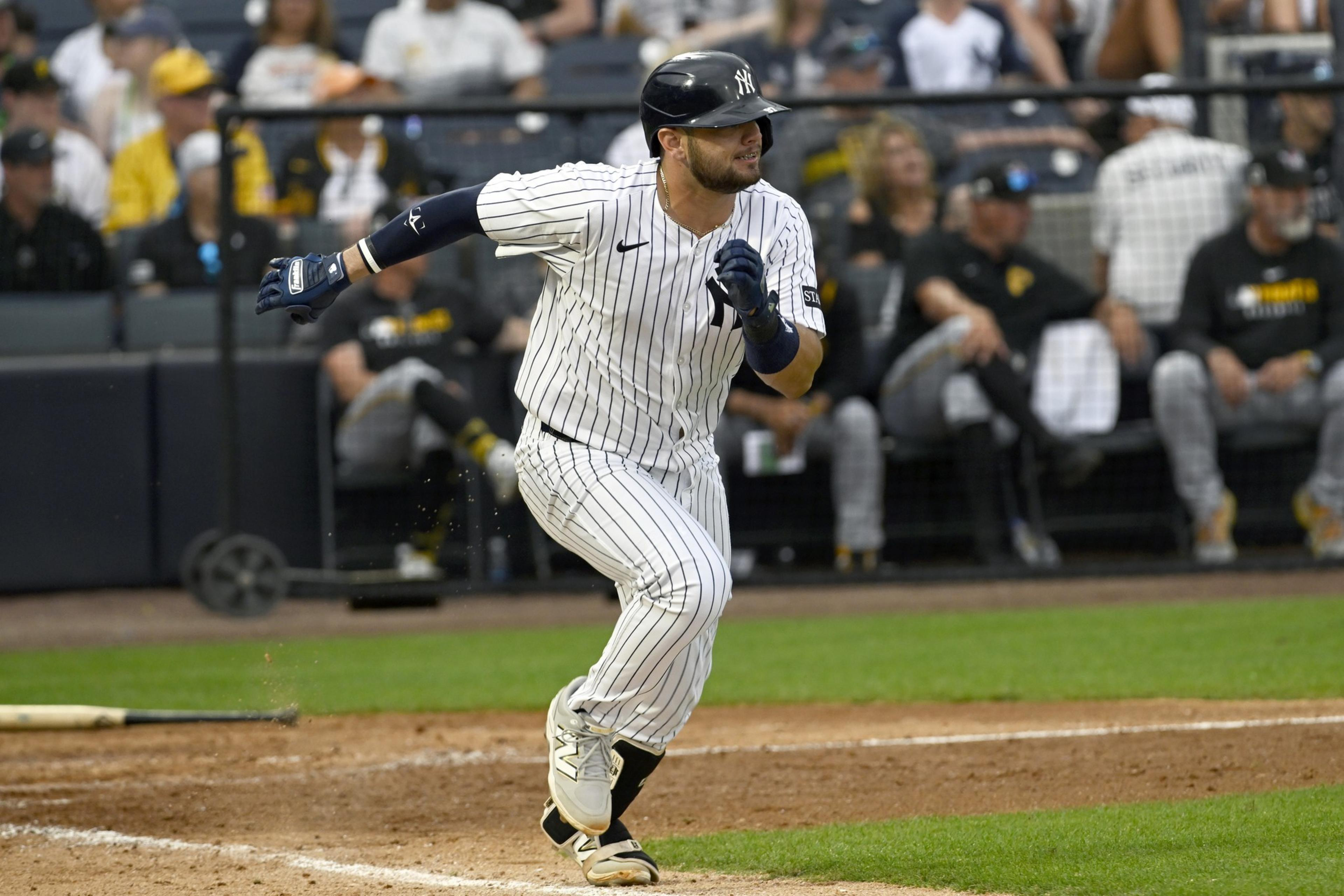 A baseball player in a New York Yankees uniform is running toward first base after hitting the ball, with blurred spectators in the background.