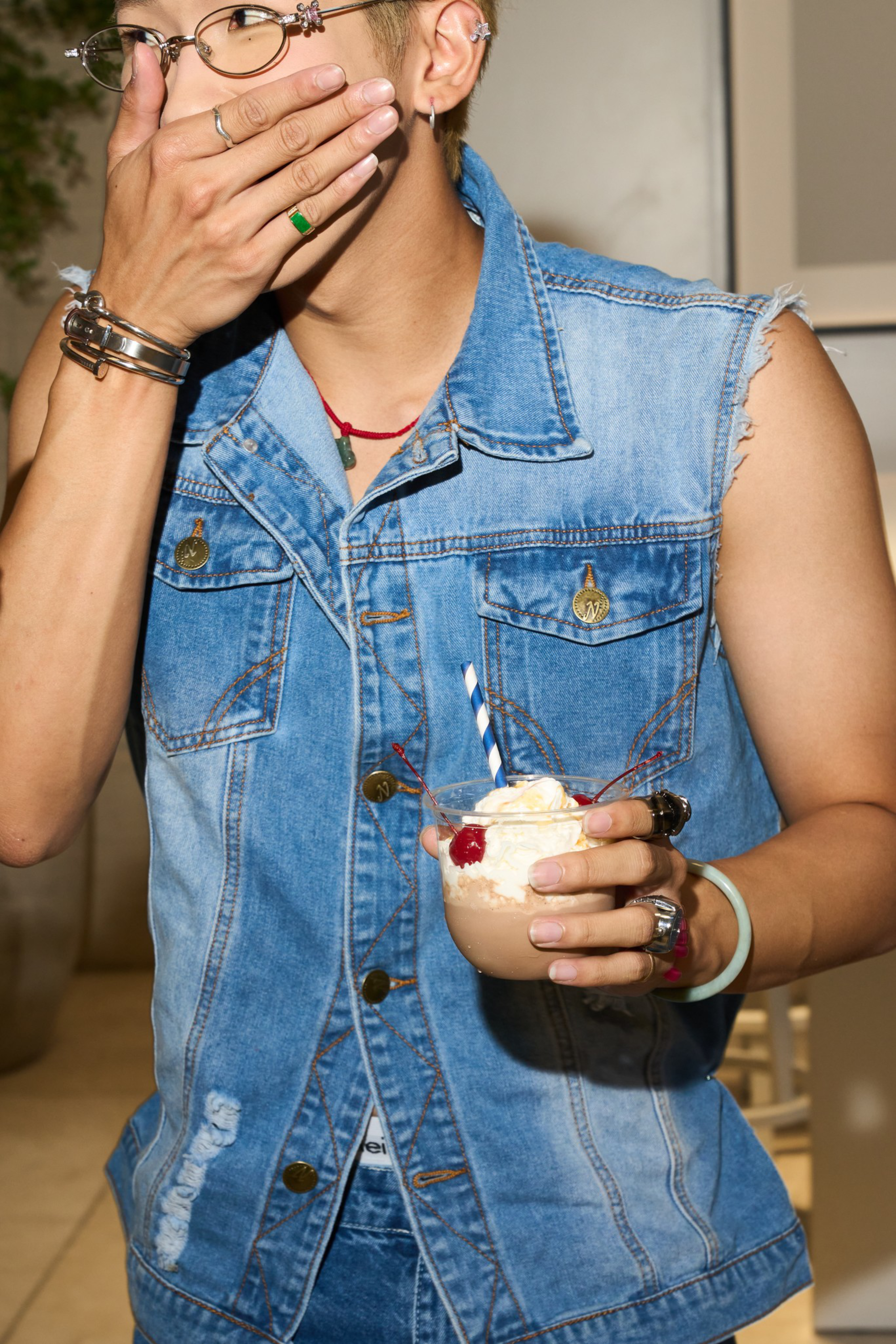 A person wearing a sleeveless denim jacket holds a whipped cream-topped drink with a cherry, straw, and various rings and bracelets, covering their mouth.