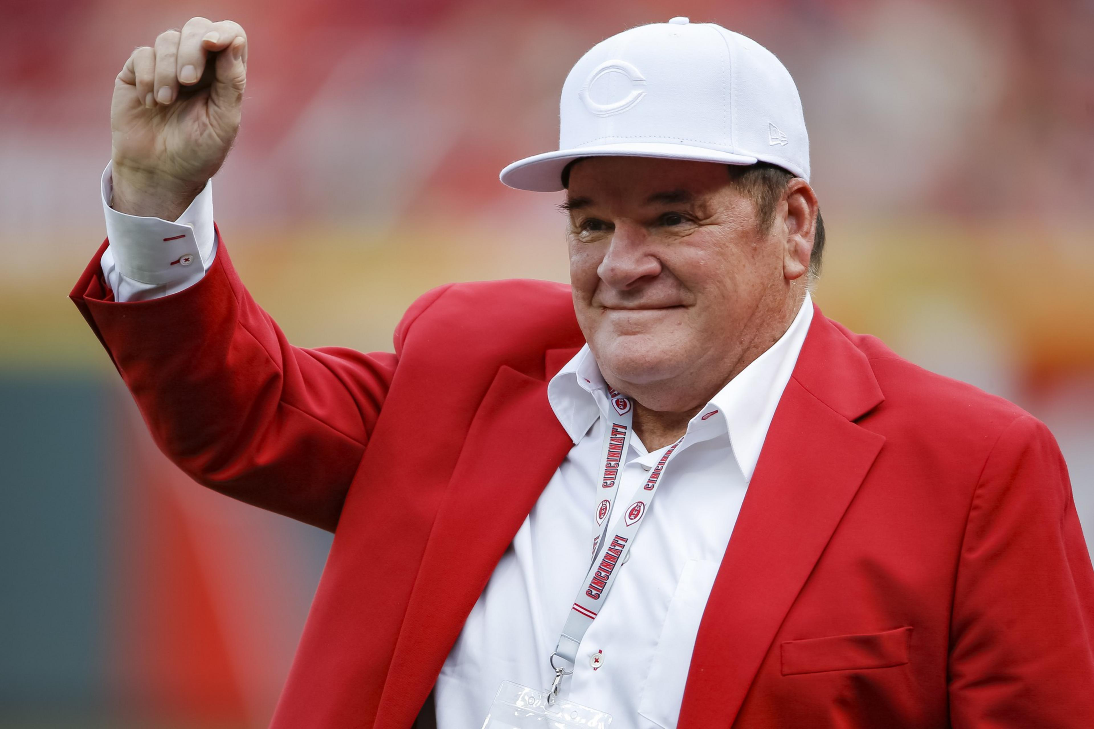 A person is wearing a bright red blazer, a white shirt, and a white baseball cap. They are smiling and raising a fist, with a lanyard around their neck.