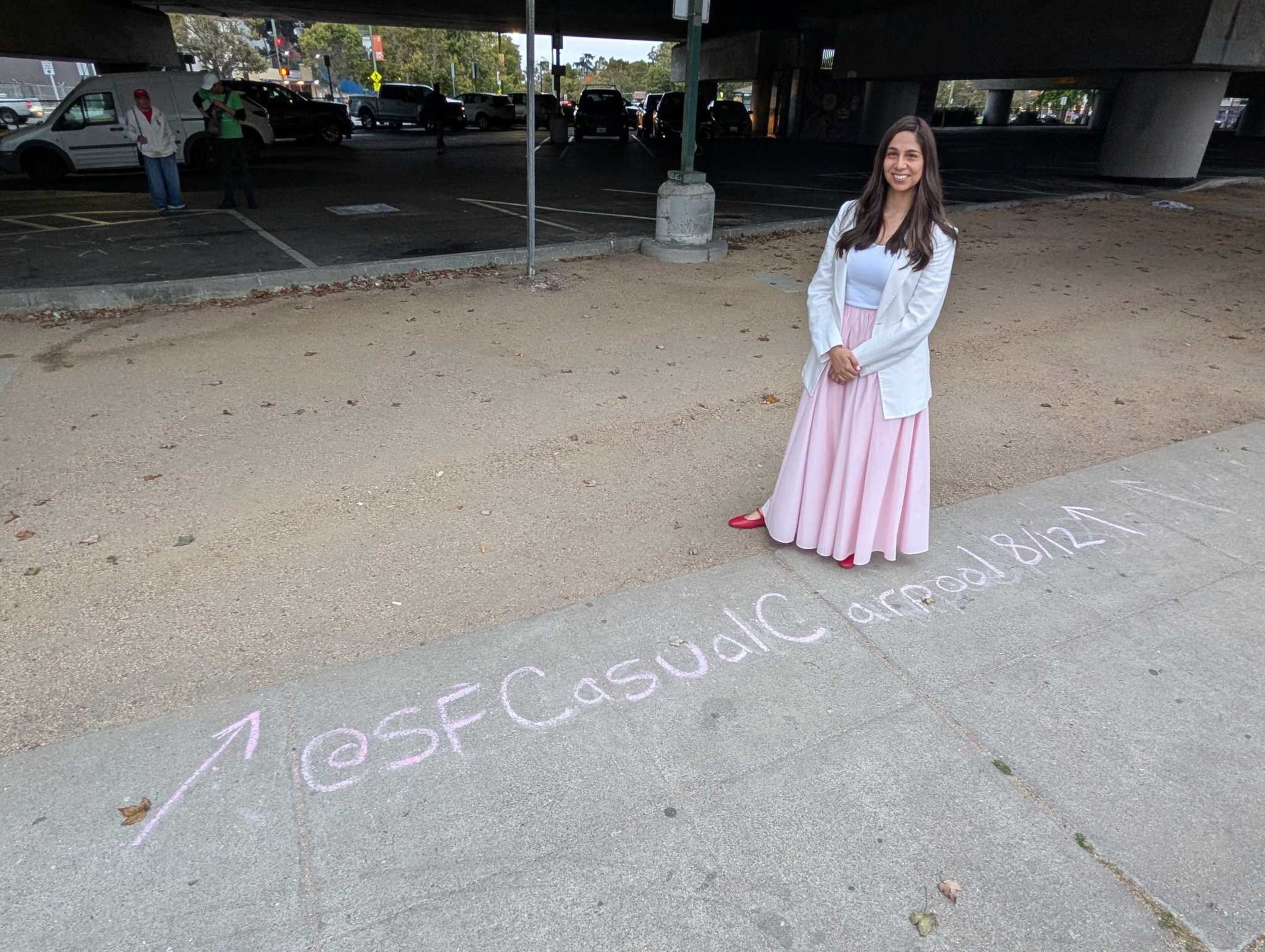 A woman in a white jacket and pink skirt stands on a sidewalk next to chalk writing that reads "@SFCasualC apparel 8/121" with an arrow pointing left.