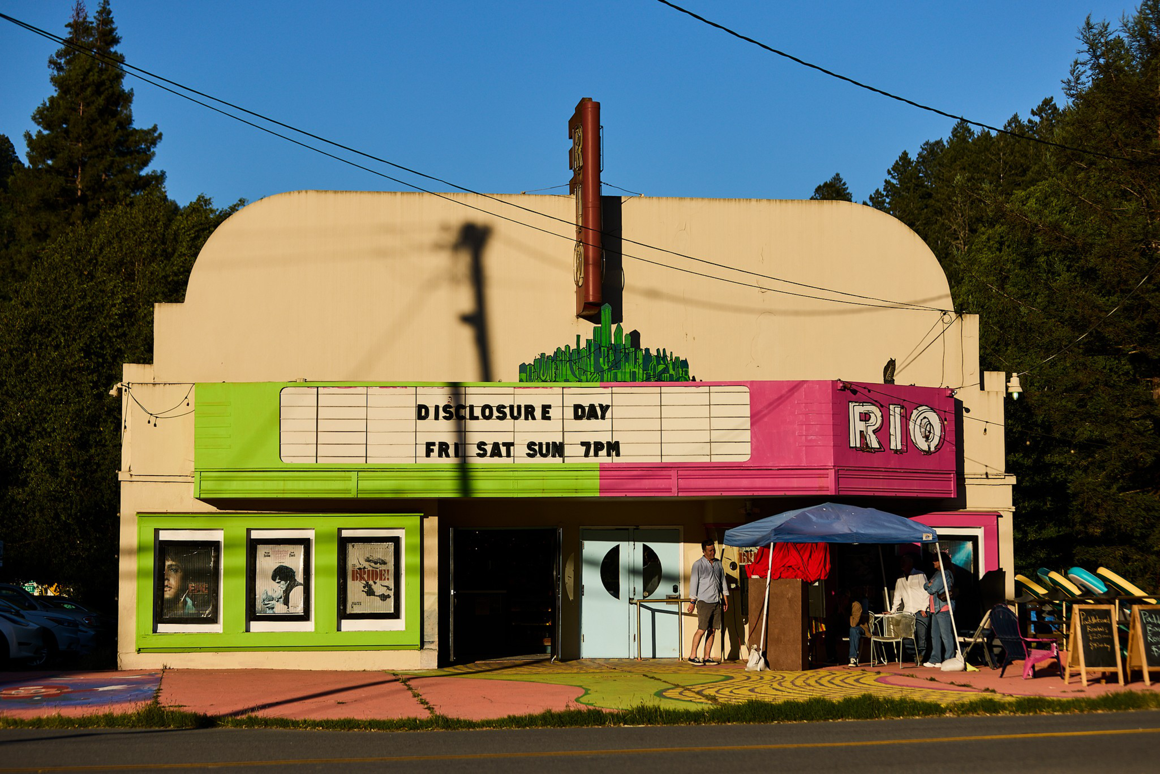 An old-fashioned movie theater with a pink and green facade displays “Disclosure Day Fri Sat Sun 7PM” and has people setting up a tent outside.