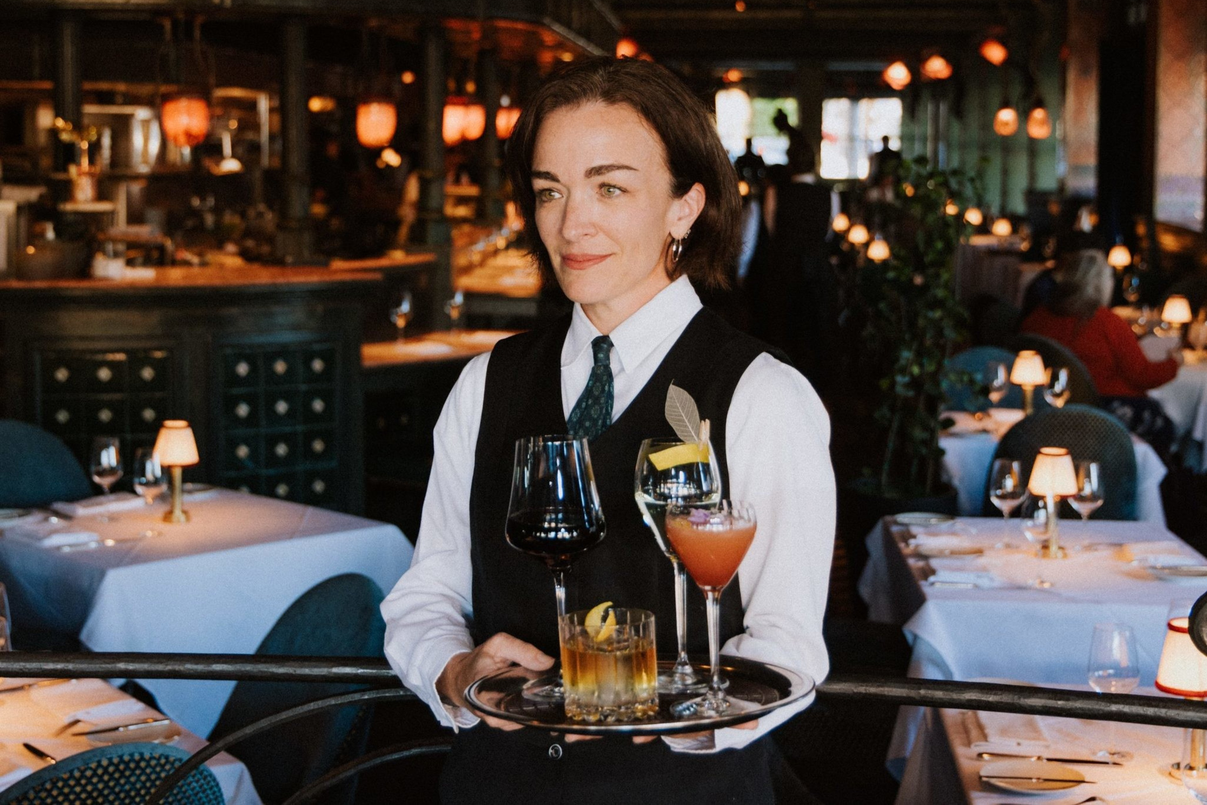 A server in a stylish restaurant carries a tray with various drinks, including wine and cocktails, amidst a dimly lit, elegant dining area with white tablecloths.