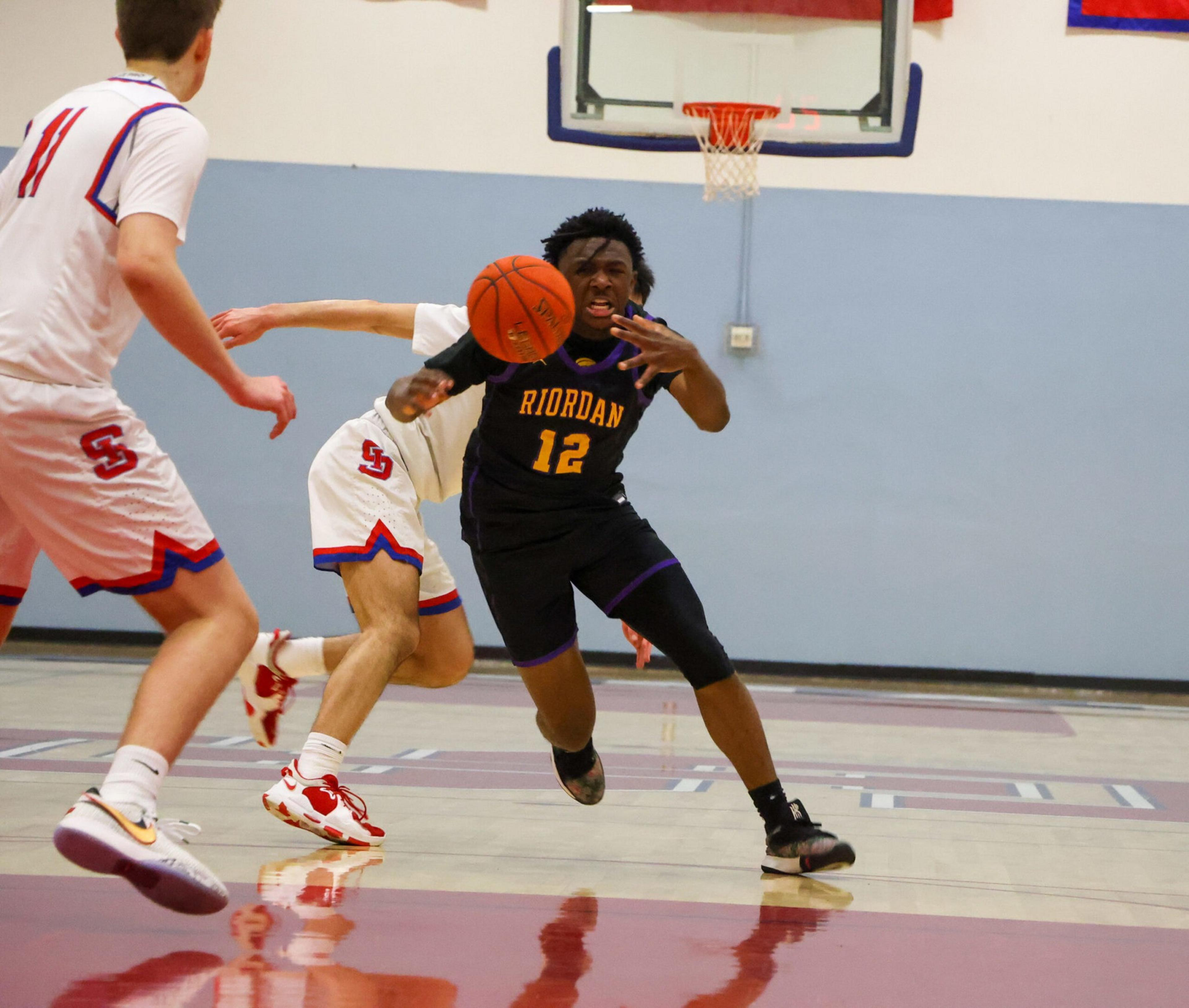 A basketball player wearing a black "Riordan" jersey lunges forward reaching for the ball while two opposing players in white jerseys defend near the hoop.
