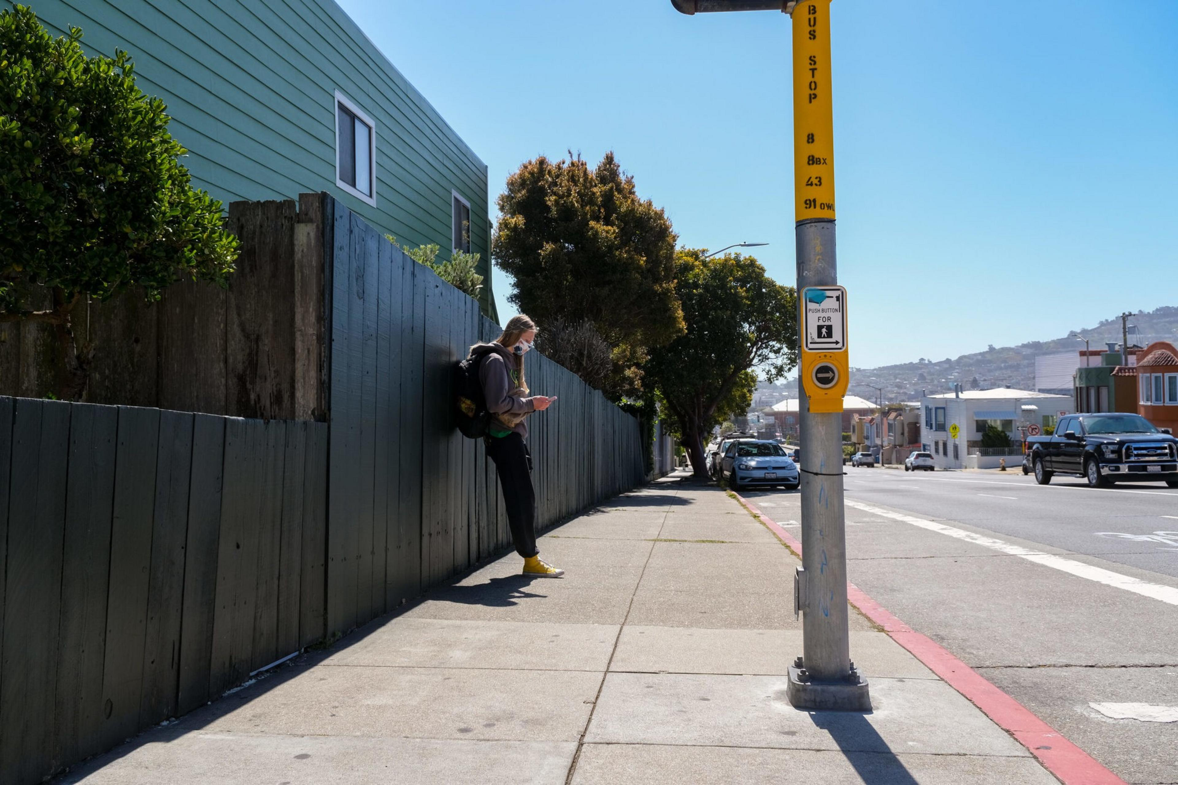 A person wearing a backpack and yellow shoes stands by a wooden fence at a bus stop on a sunny sidewalk beside a street with cars.