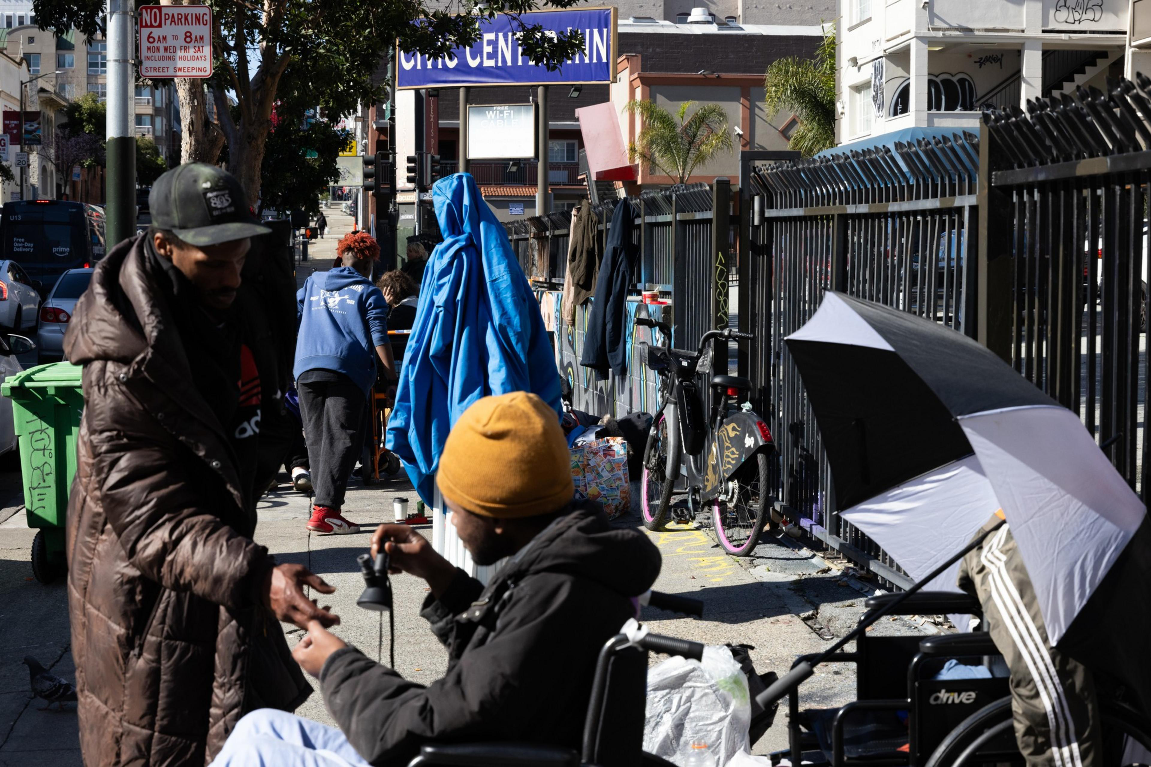 People are interacting on a busy sidewalk near a &quot;Civic Center&quot; sign. One person is sitting in a wheelchair, holding an item, while another stands nearby.