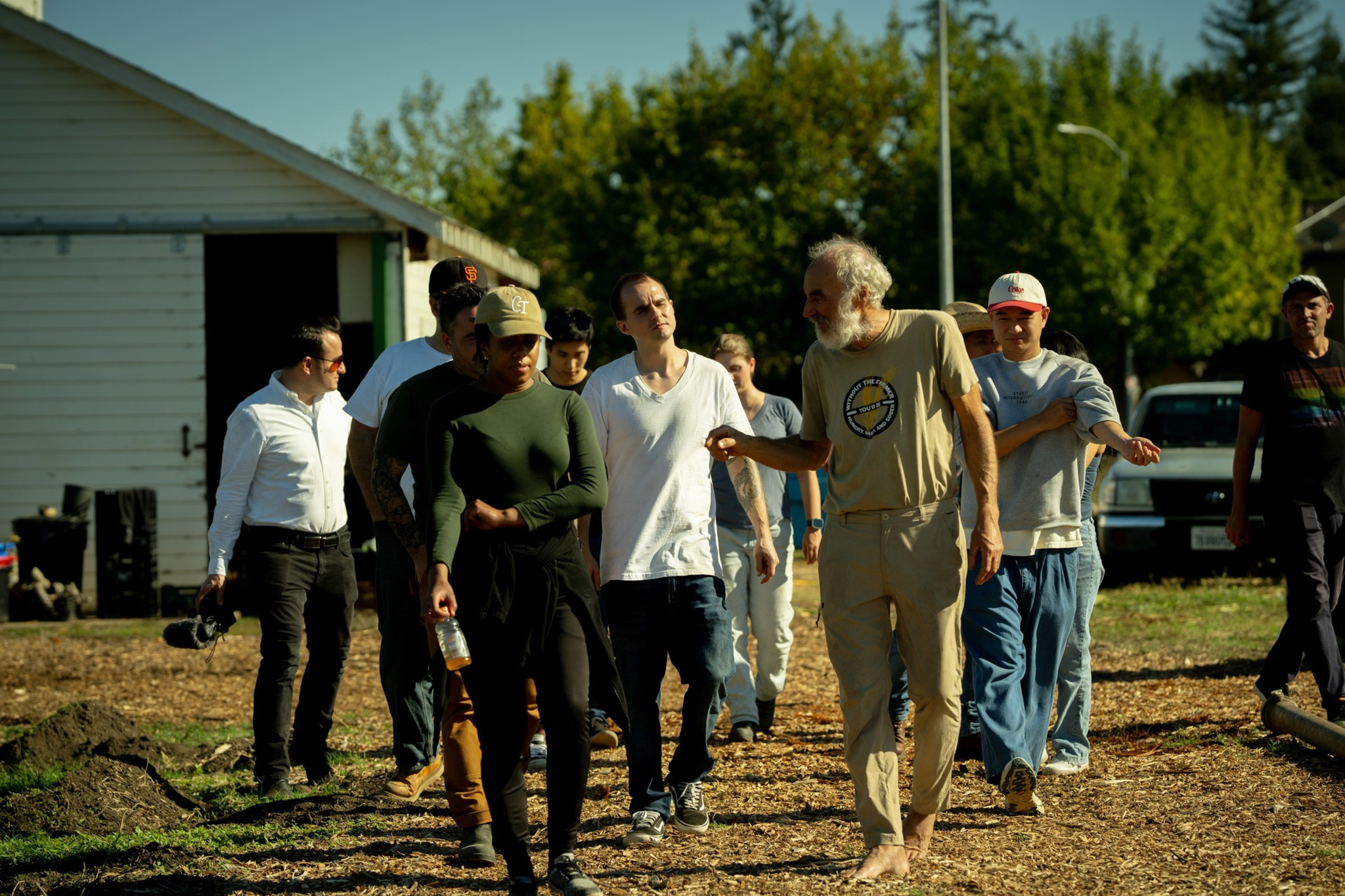 A diverse group of people walk outdoors on a sunny day, with trees and a building in the background; one man is barefoot and gesturing while talking.
