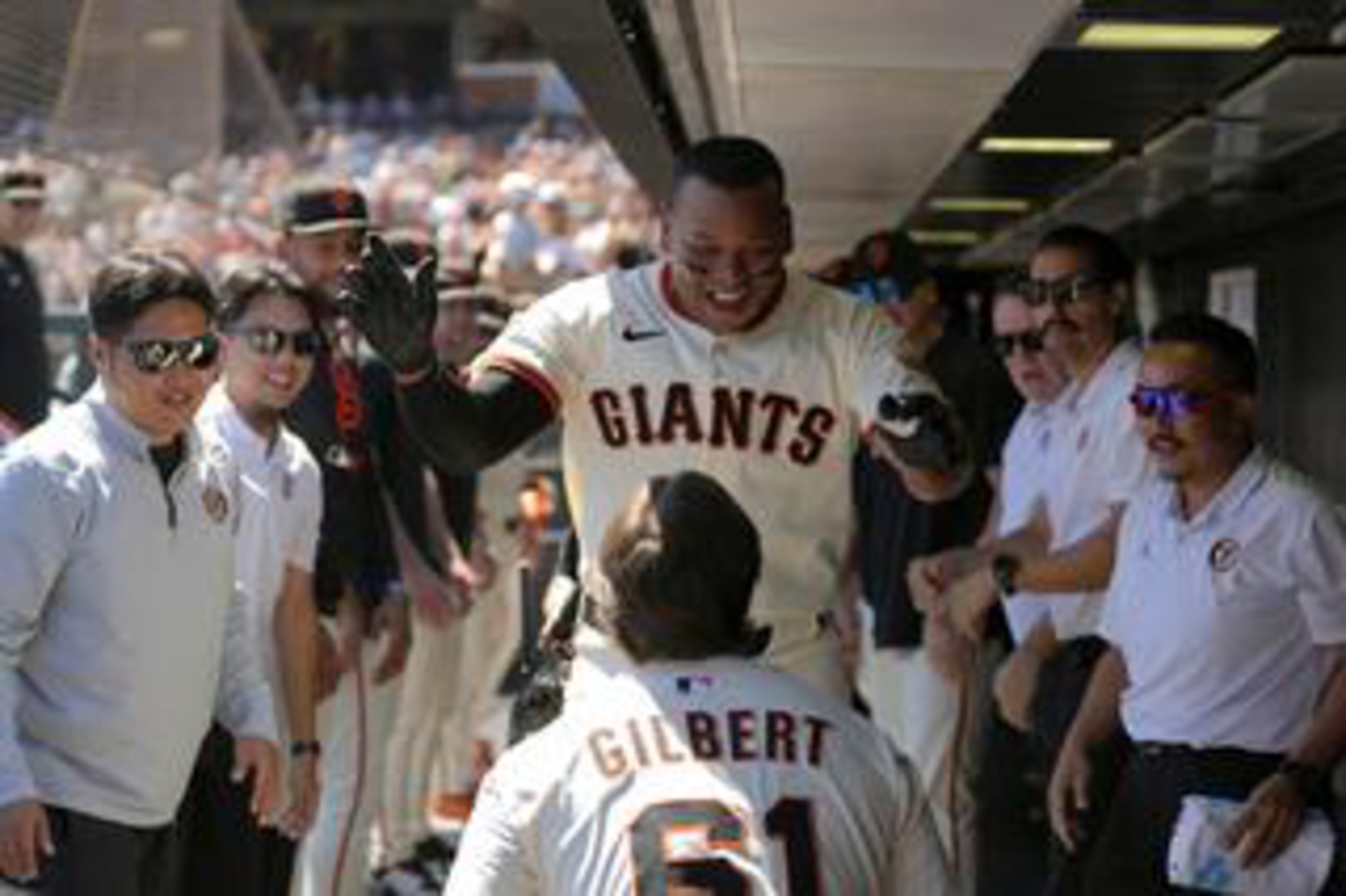 A Giants player smiles and raises his hands to high-five teammate Gilbert, while staff and players cheer in a baseball dugout.