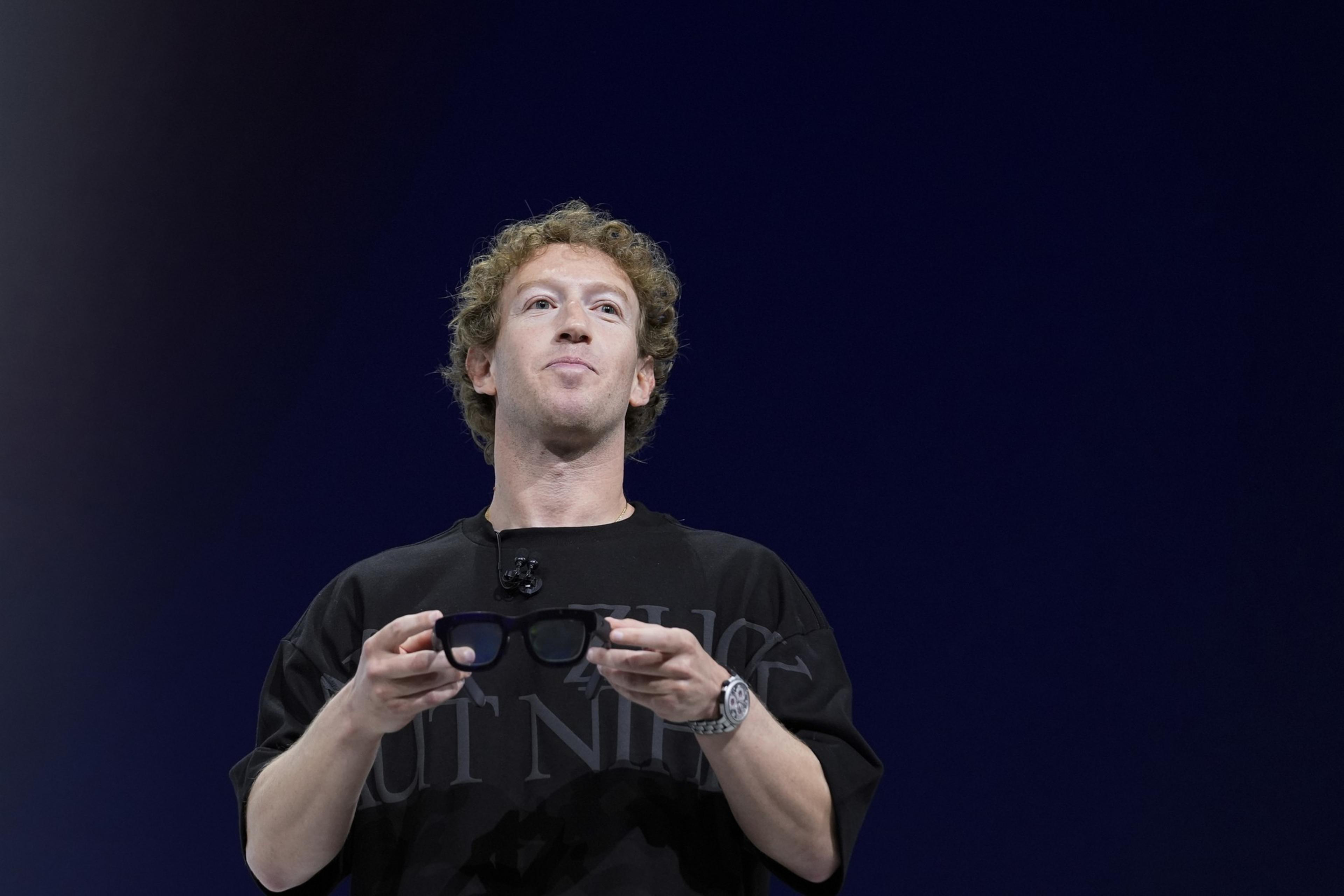 A person with curly hair is holding smart glasses, wearing a black shirt, and speaking on stage with a microphone clipped to their shirt against a dark background.