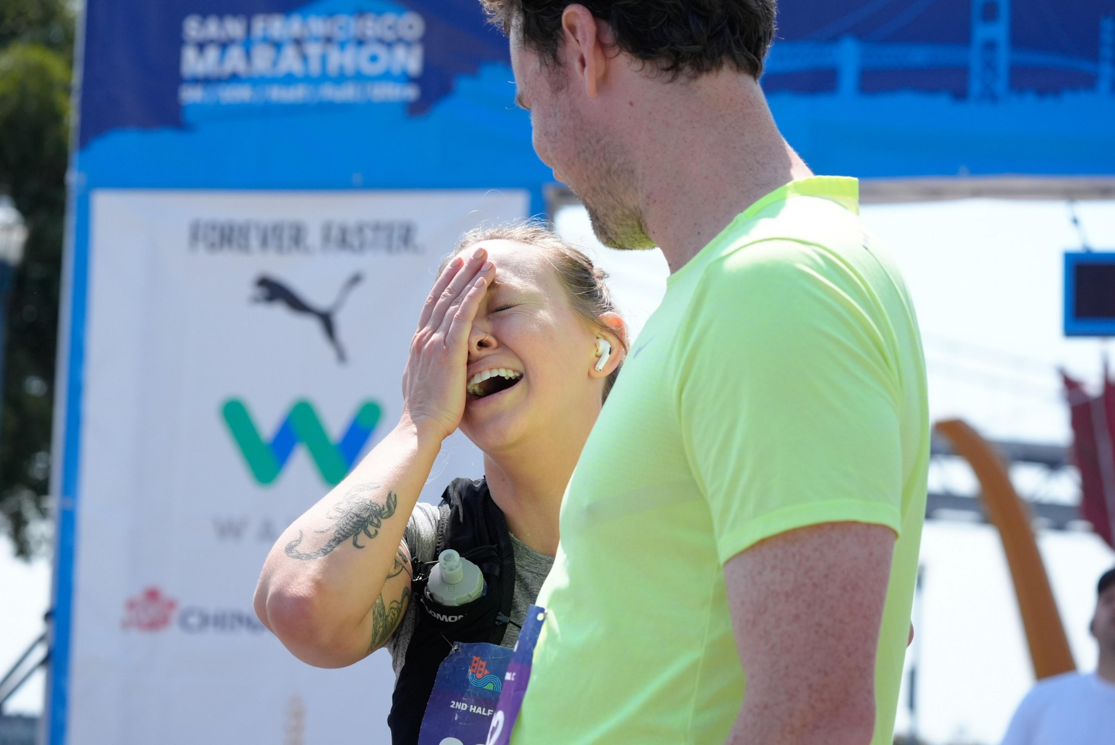 A woman laughs while covering her face, standing beside a man in a neon yellow shirt at the San Francisco Marathon finish line, with a blue marathon banner in the background.