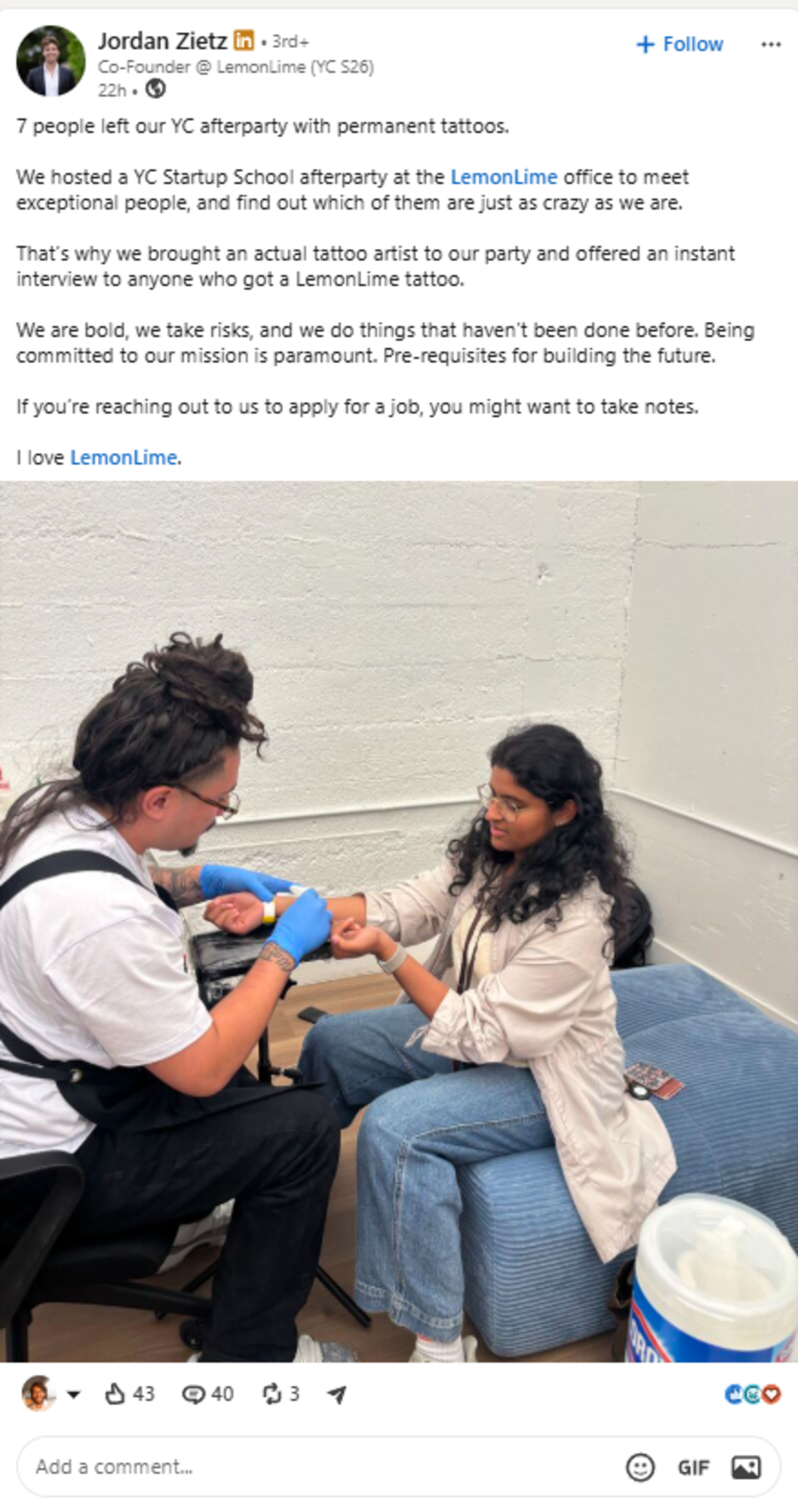 A tattoo artist wearing blue gloves tattoos a woman’s inner wrist as she sits on a blue ottoman in a bright, minimalist room.