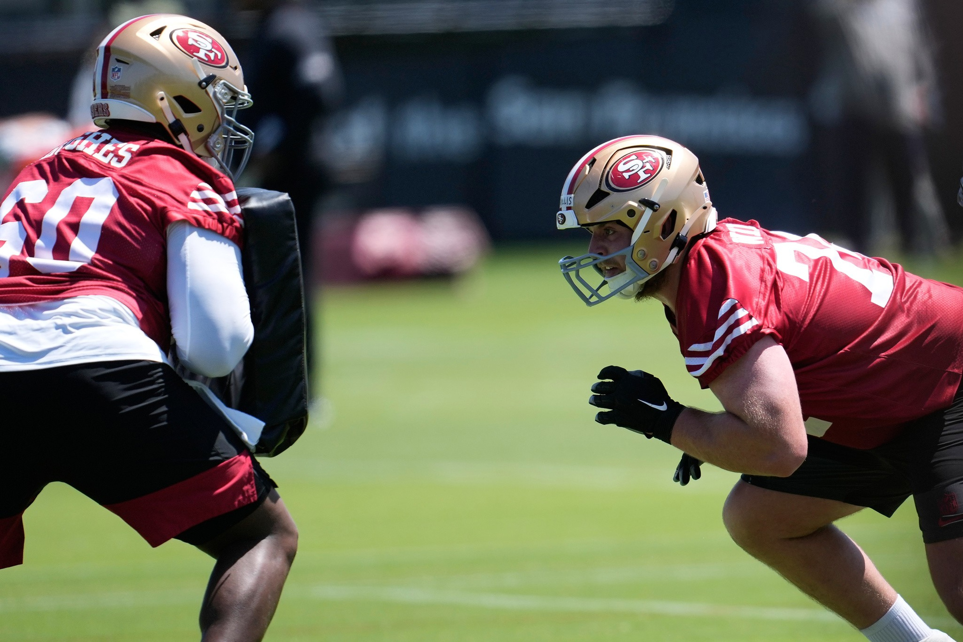 Two San Francisco 49ers players in helmets and red jerseys practice on a grassy field, with one in a crouched position facing a blocking pad.