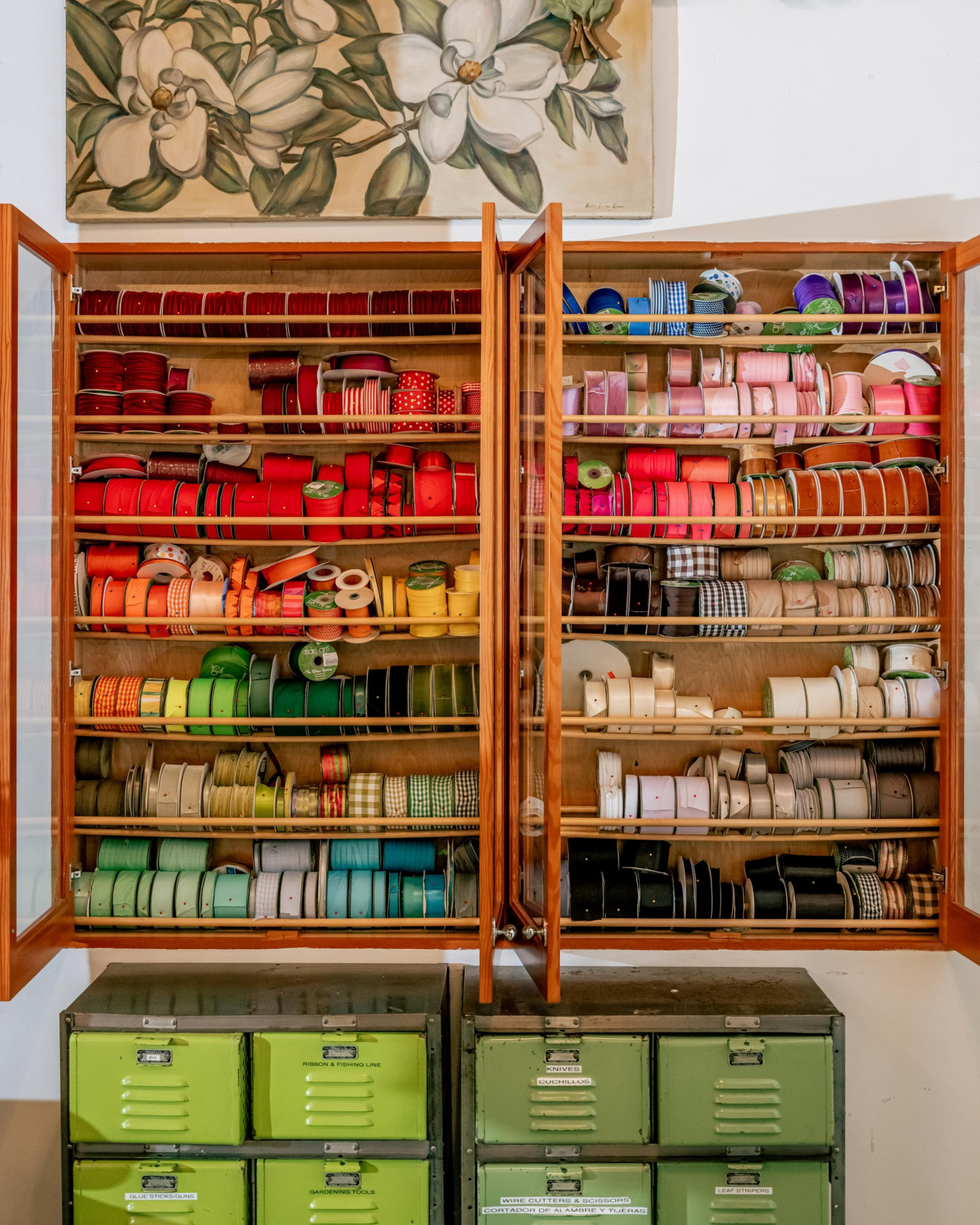 Wooden cabinet with glass doors holds numerous colorful ribbon rolls organized by color, above stacked green metal storage drawers, under a floral painting.
