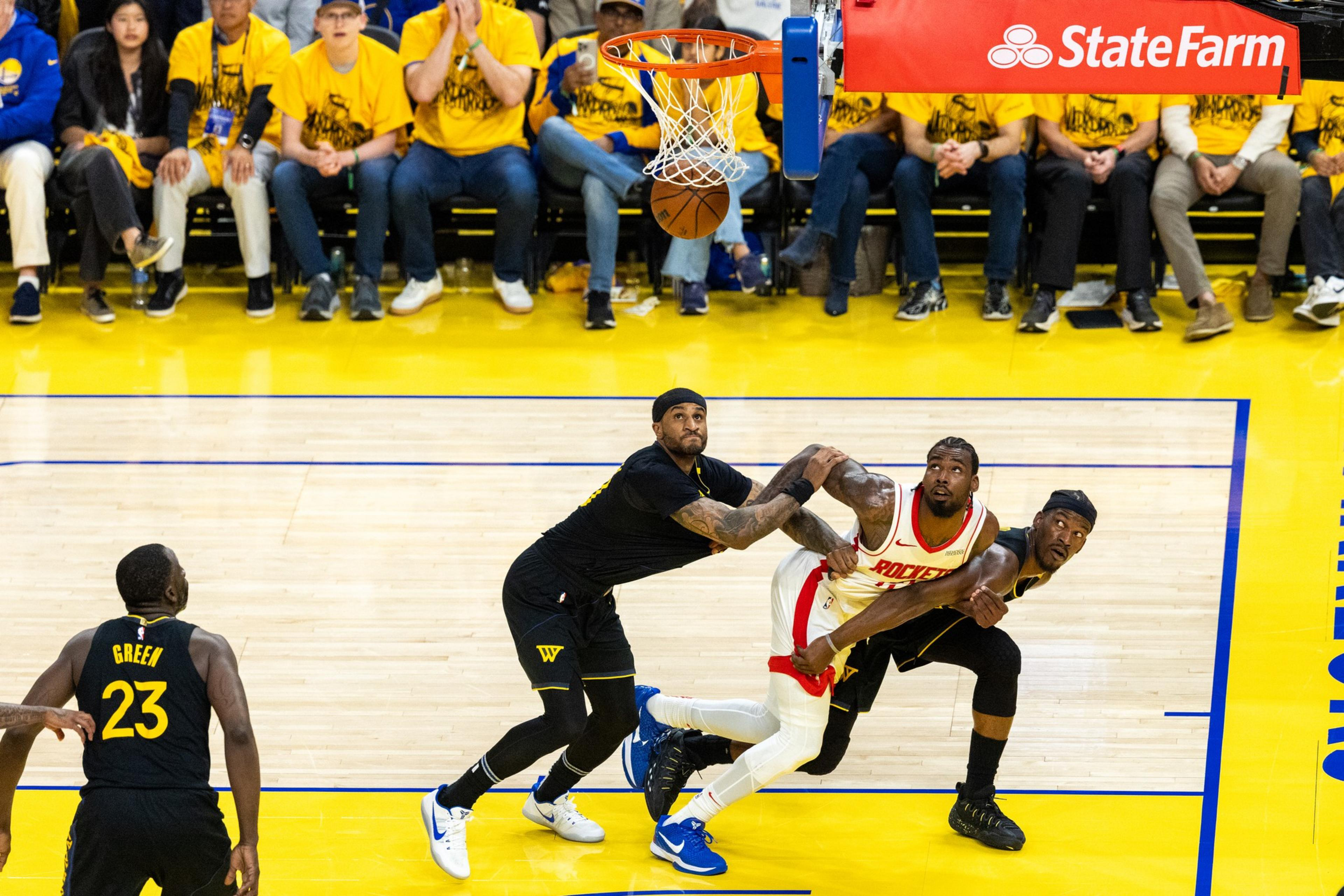 A basketball game is in action with three players near the hoop. Fans in yellow shirts fill the stands, and the ball is about to enter the basket.