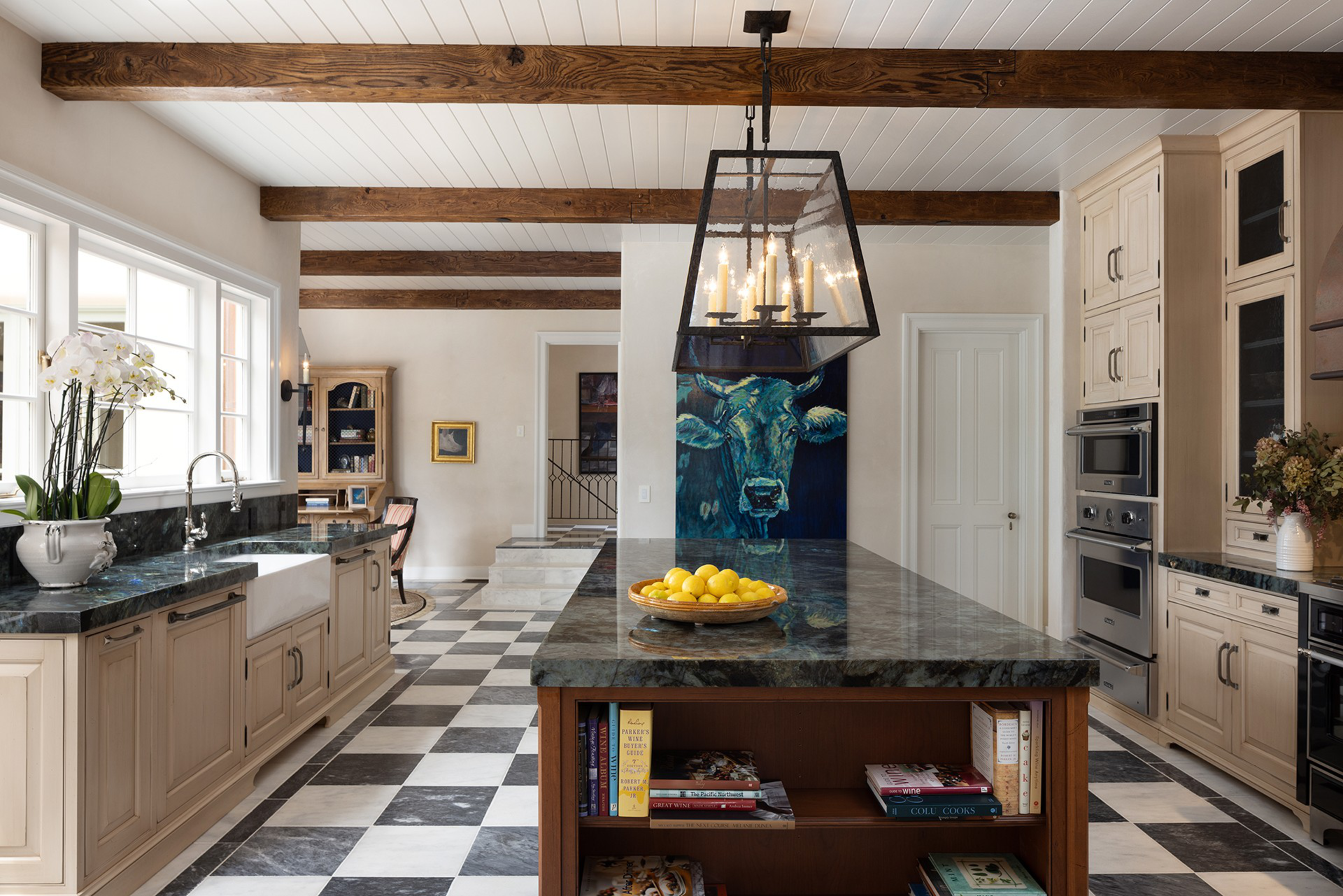 A spacious kitchen features black-and-white checkered floors, wooden beams on a white ceiling, a central island with a bowl of lemons, and a blue cow painting.