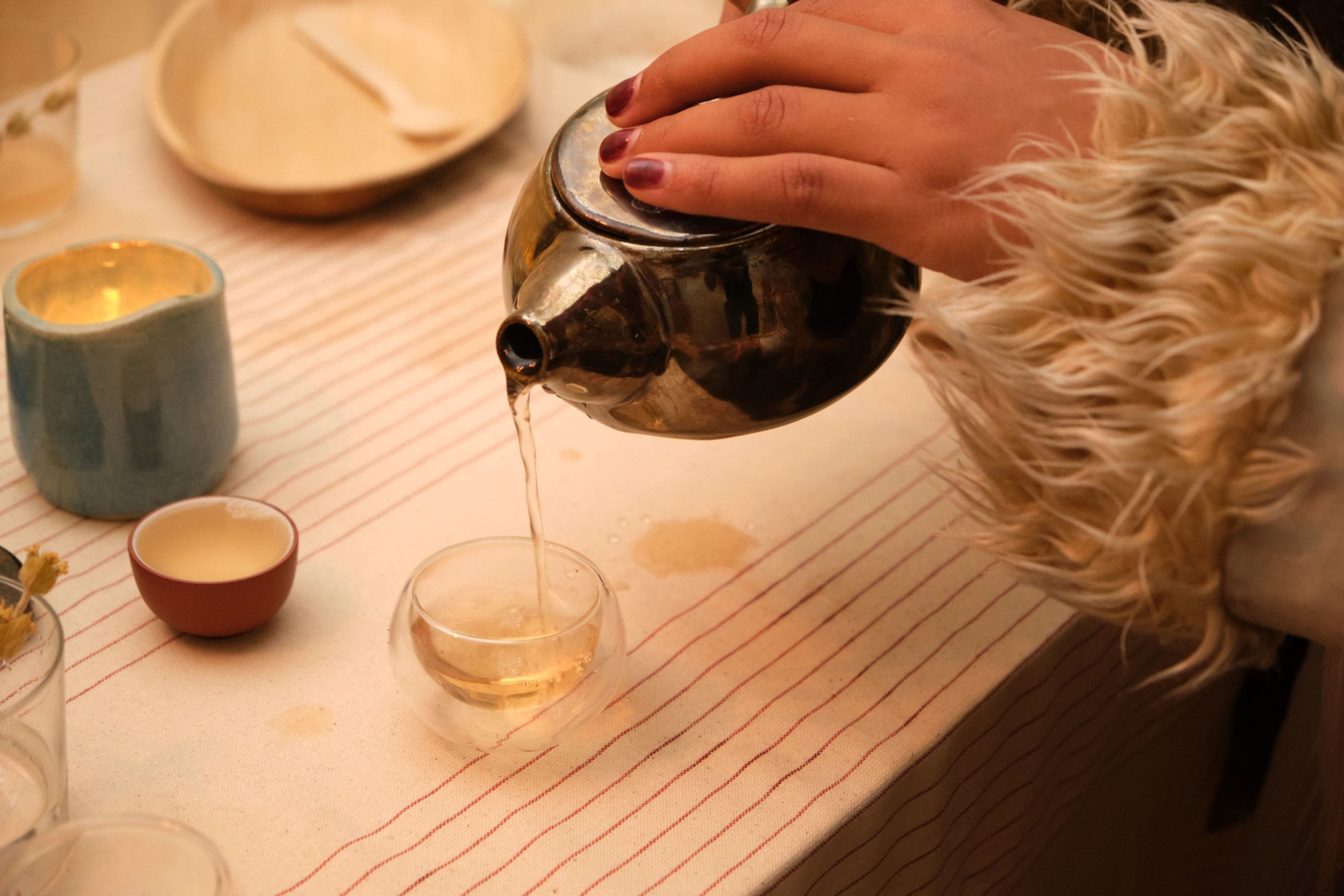 A hand with red nail polish and a furry sleeve pours tea from a metal teapot into a small glass cup on a striped tablecloth.