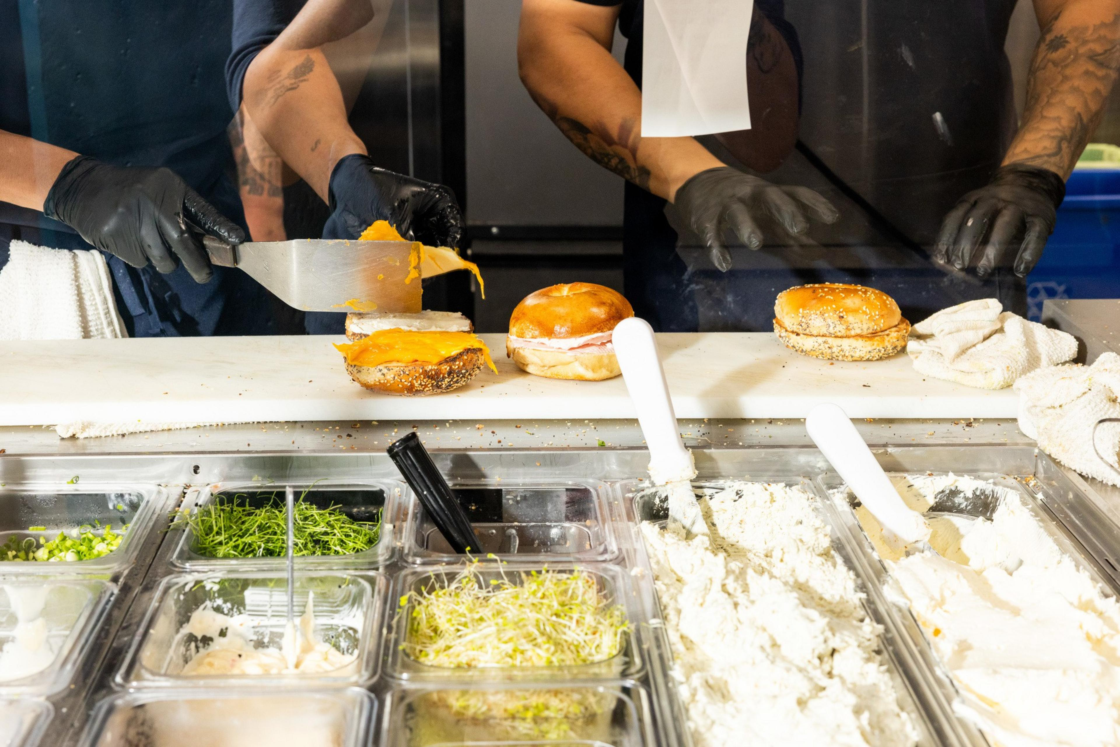 A person wearing black gloves is placing a slice of melted cheese on a sandwich; various sandwich toppings are visible below on the counter.