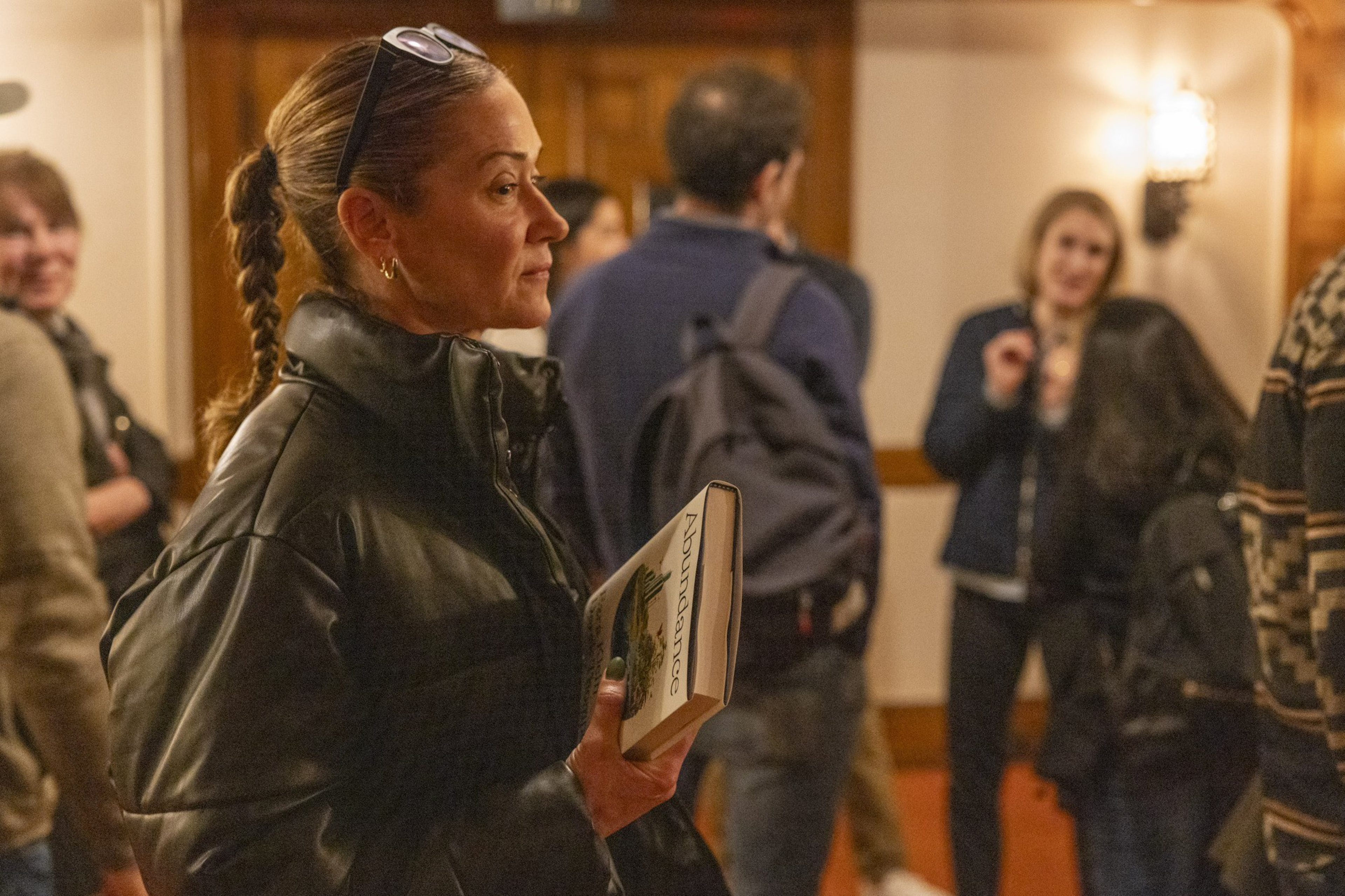 A woman with braided hair and sunglasses on her head holds a book titled &quot;Abundance&quot; while standing in a room with several people engaged in conversation.