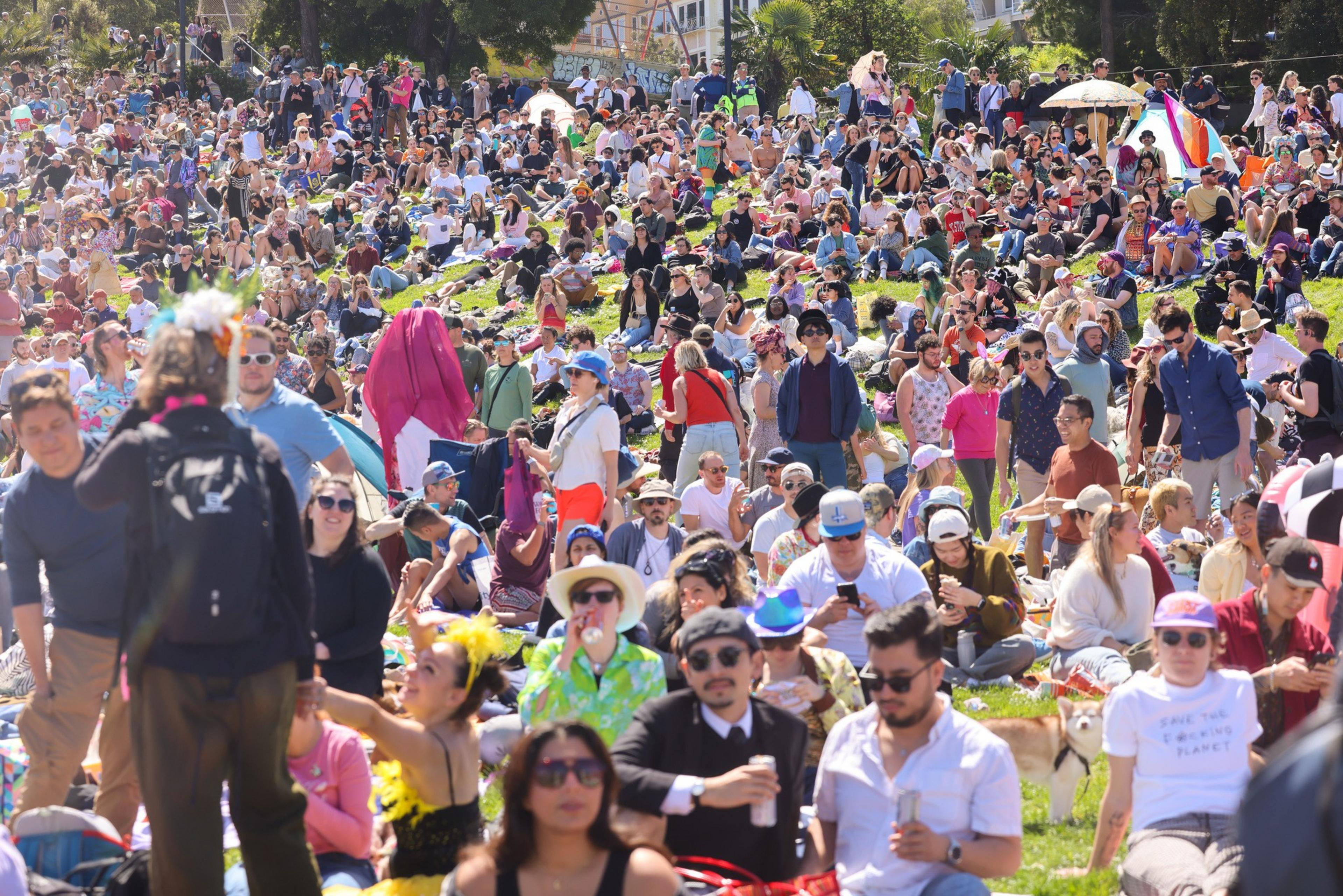 A crowded park with many people sitting on grass, enjoying a sunny day outdoors.
