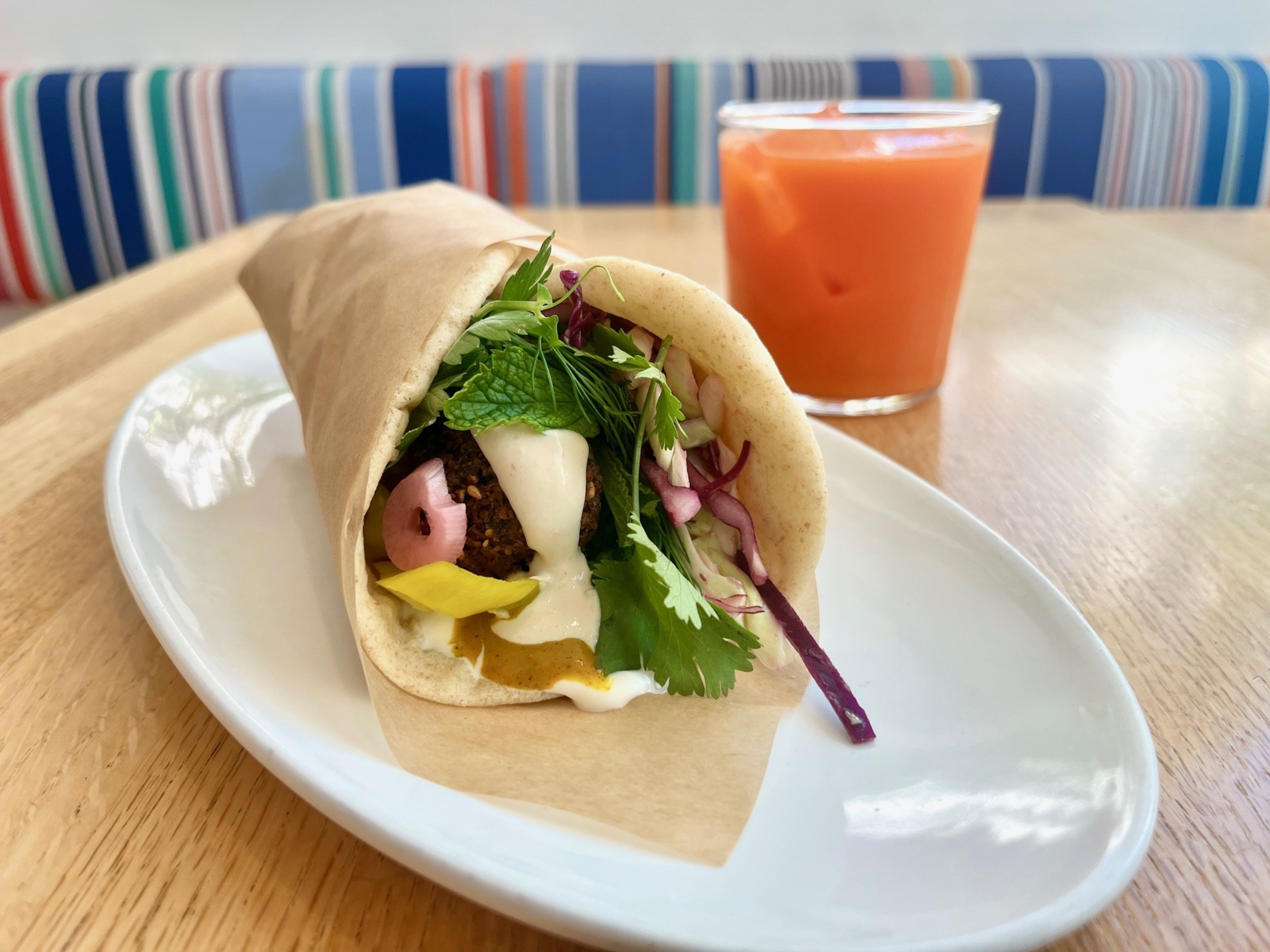 A falafel wrap filled with vegetables and sauce is on a white plate, with a glass of orange juice in the background. The setting is a colorful, striped booth.