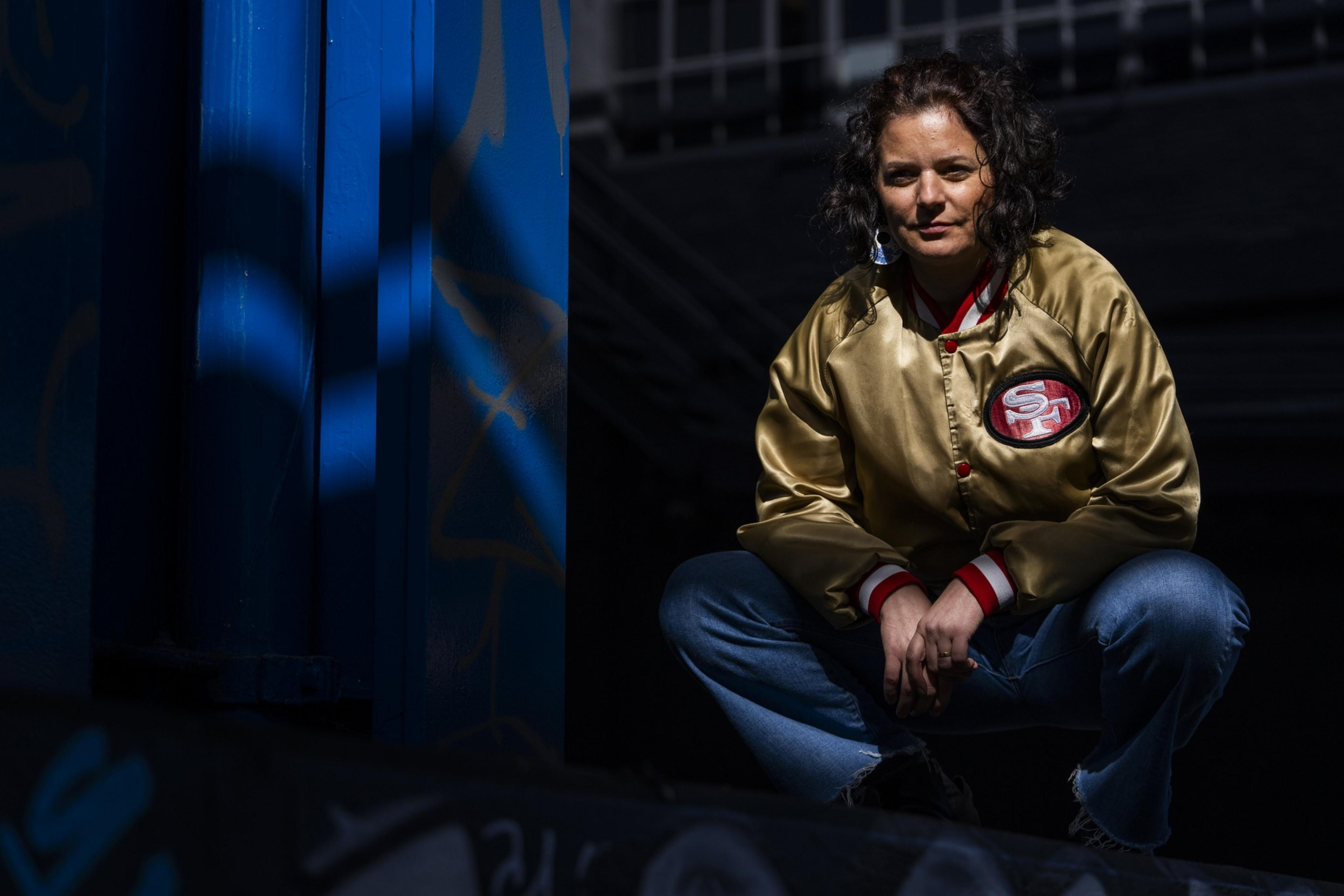 A person in a gold and red sports jacket squats in a shadowy area with graffiti, sunlit face and jacket prominent.