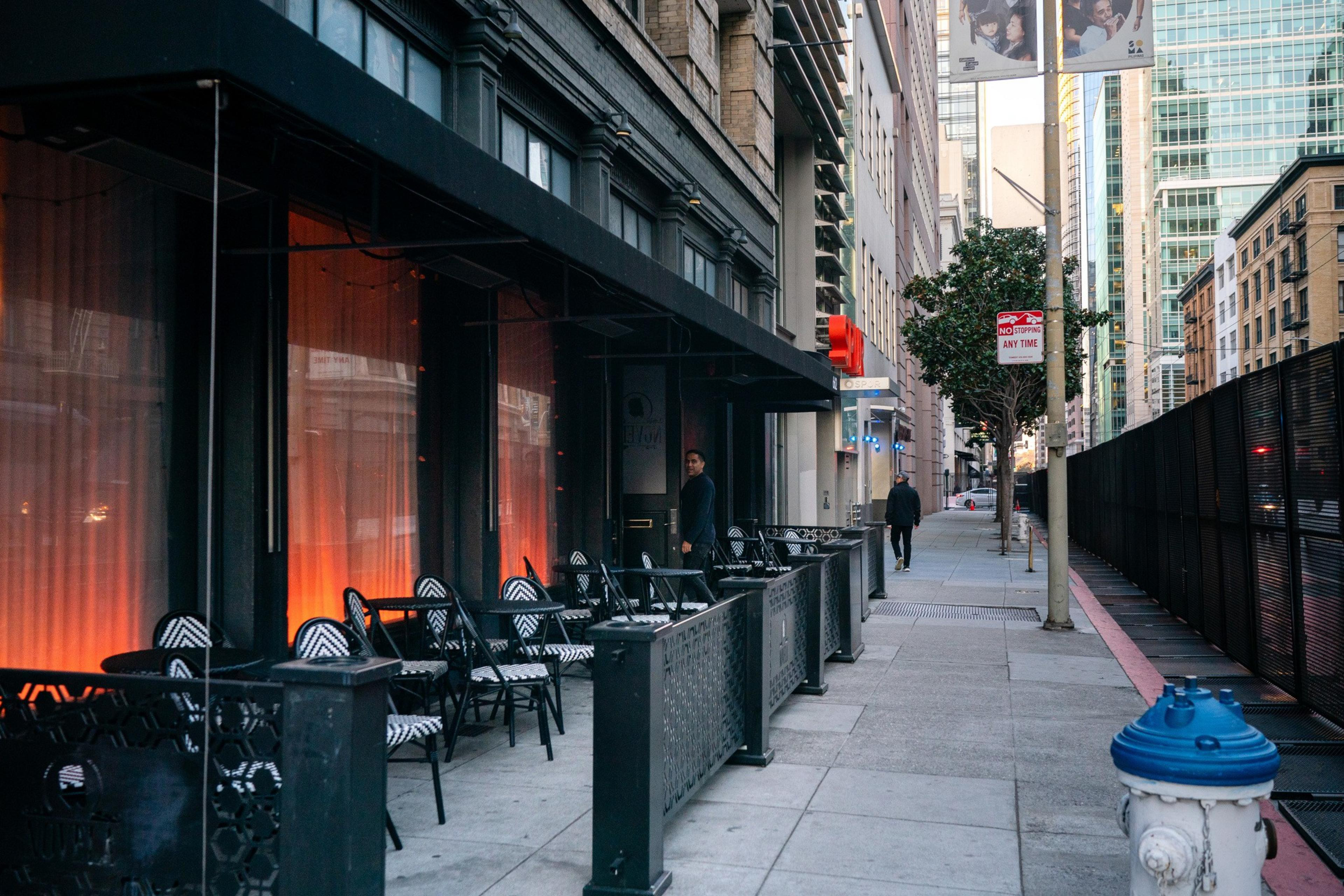 An view of an empty outdoor seating are on a city block with a tall black fence on the opposite side.