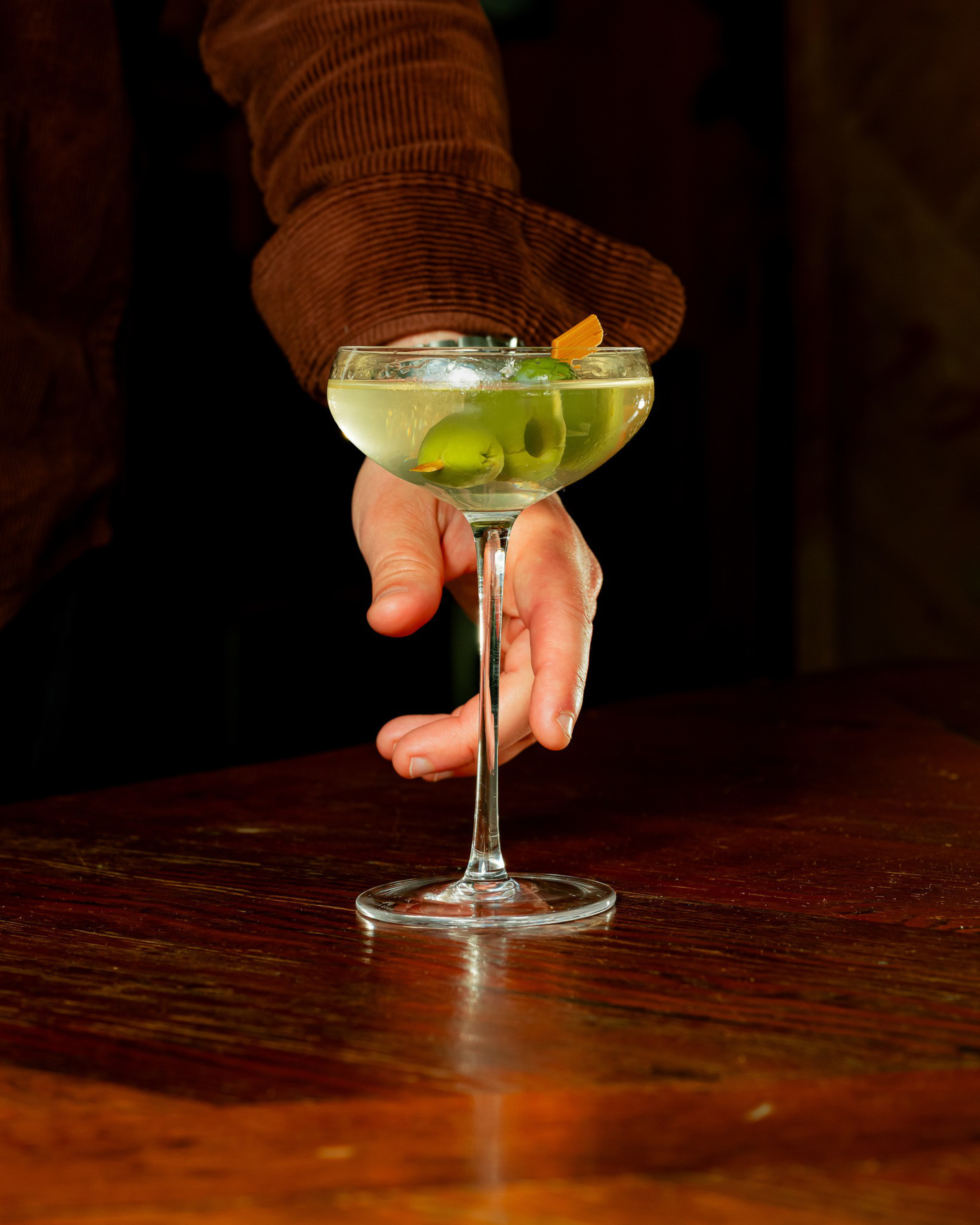 A person in a brown jacket holds a cocktail glass filled with a pale liquid and two green olives, garnished with a twist, over a wooden table.