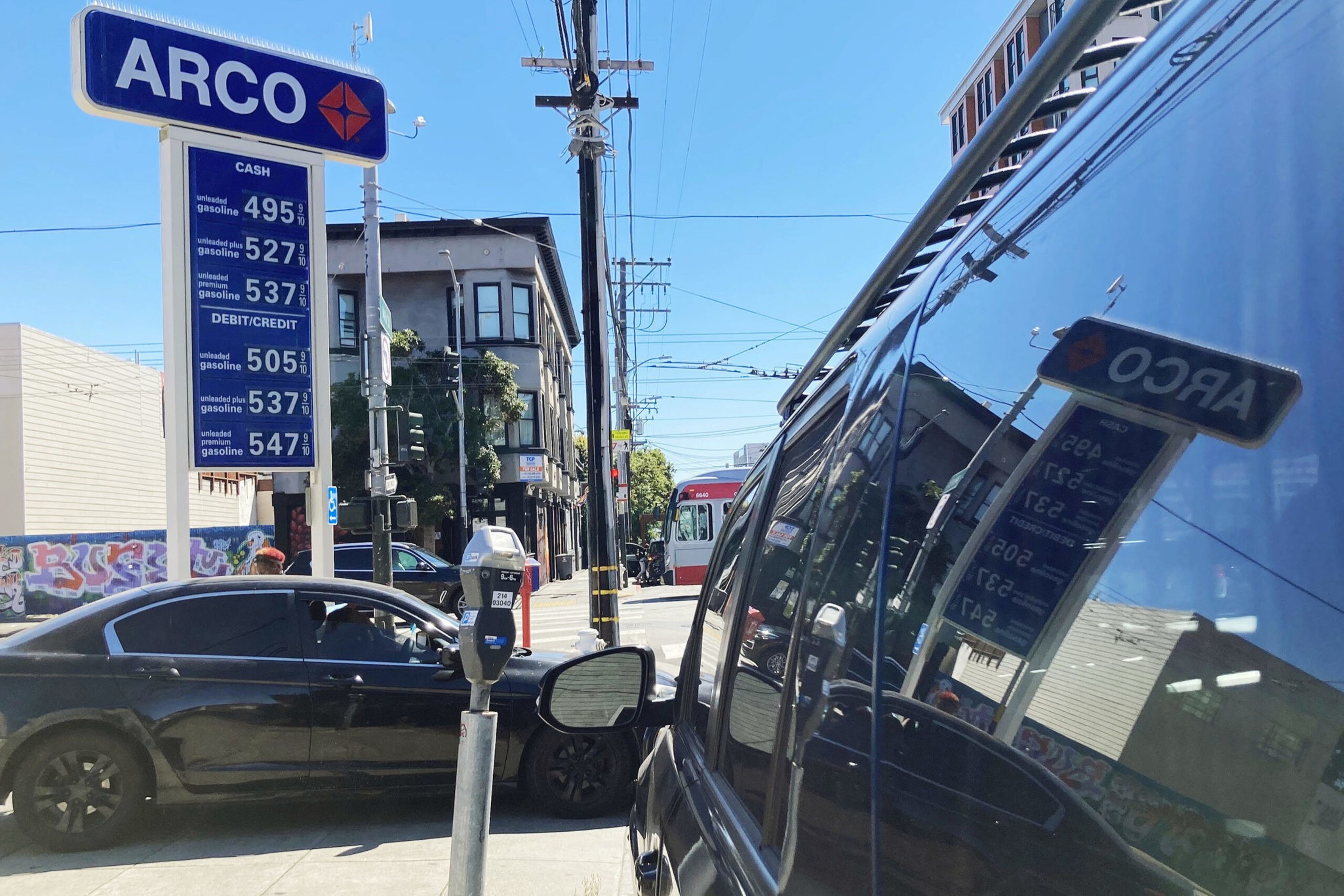 An ARCO gas station price sign lists fuel prices ranging from $4.95 to $5.47 per gallon, while cars are parked along a sunny urban street.