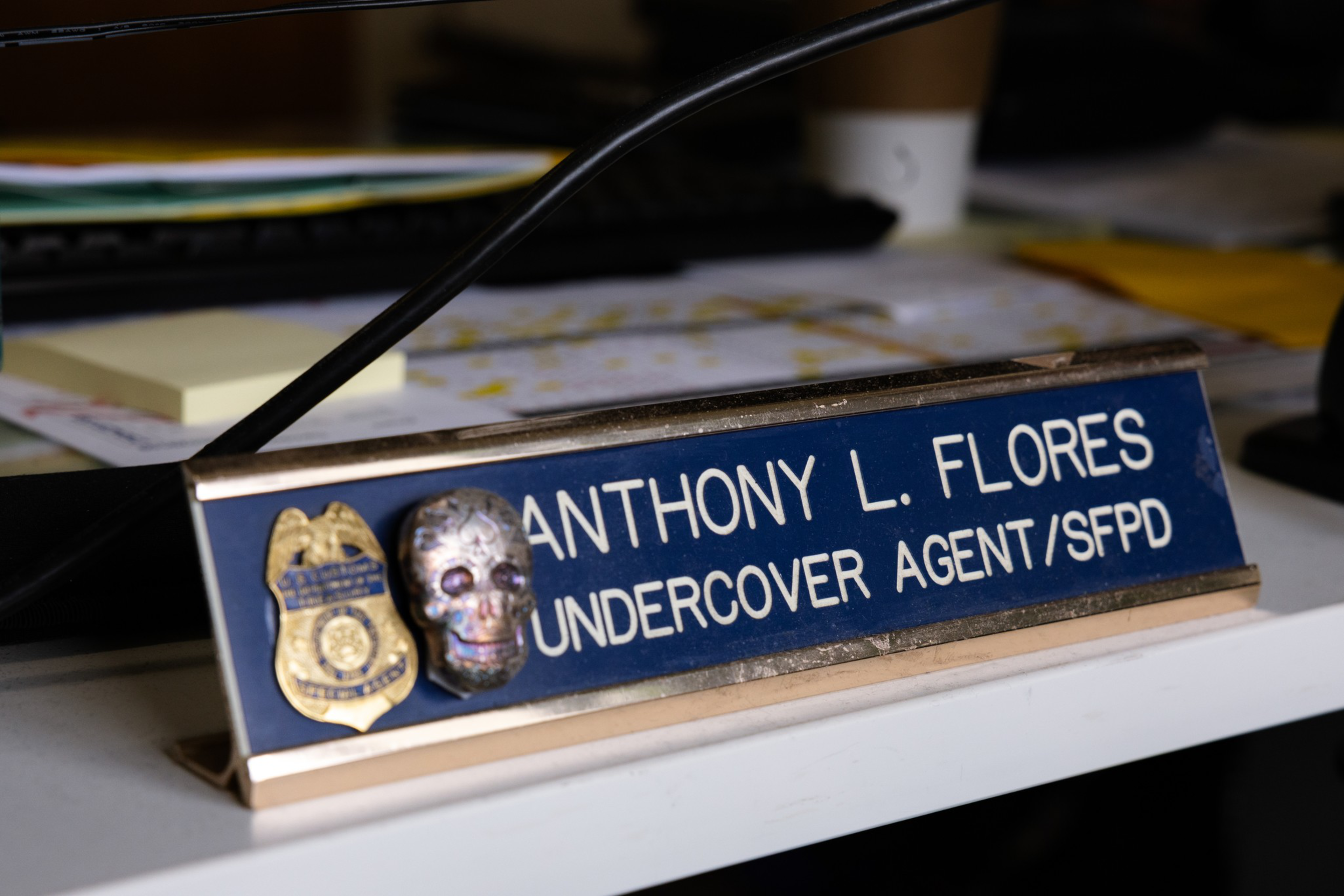 A desk nameplate with a police badge and skull figurine reads “Anthony L. Flores, Undercover Agent/SFPD,” resting on a white desk with papers behind.