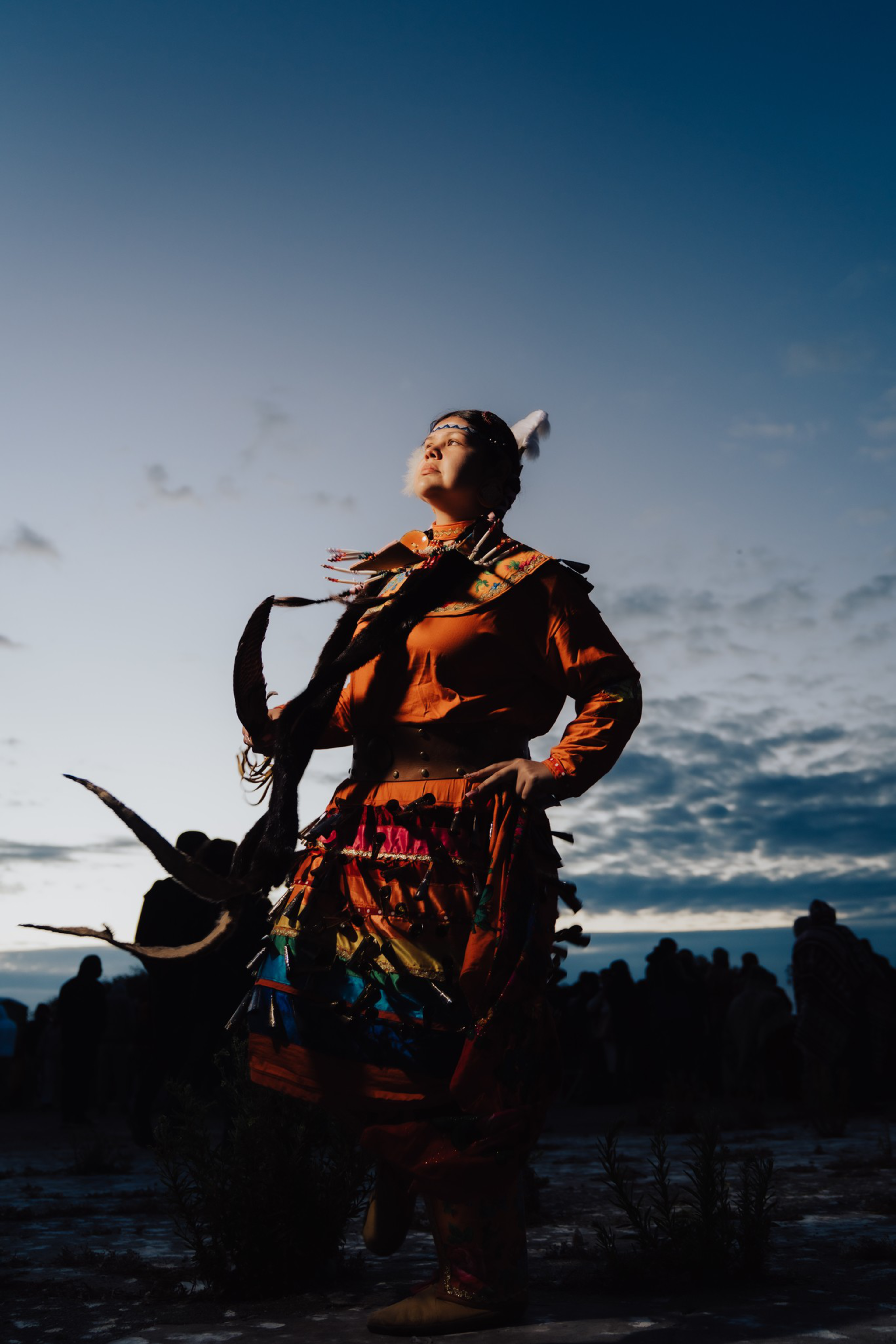 A person in vibrant traditional attire with feathers and colorful decorations poses confidently against a dusky sky, with shadowed figures in the background.