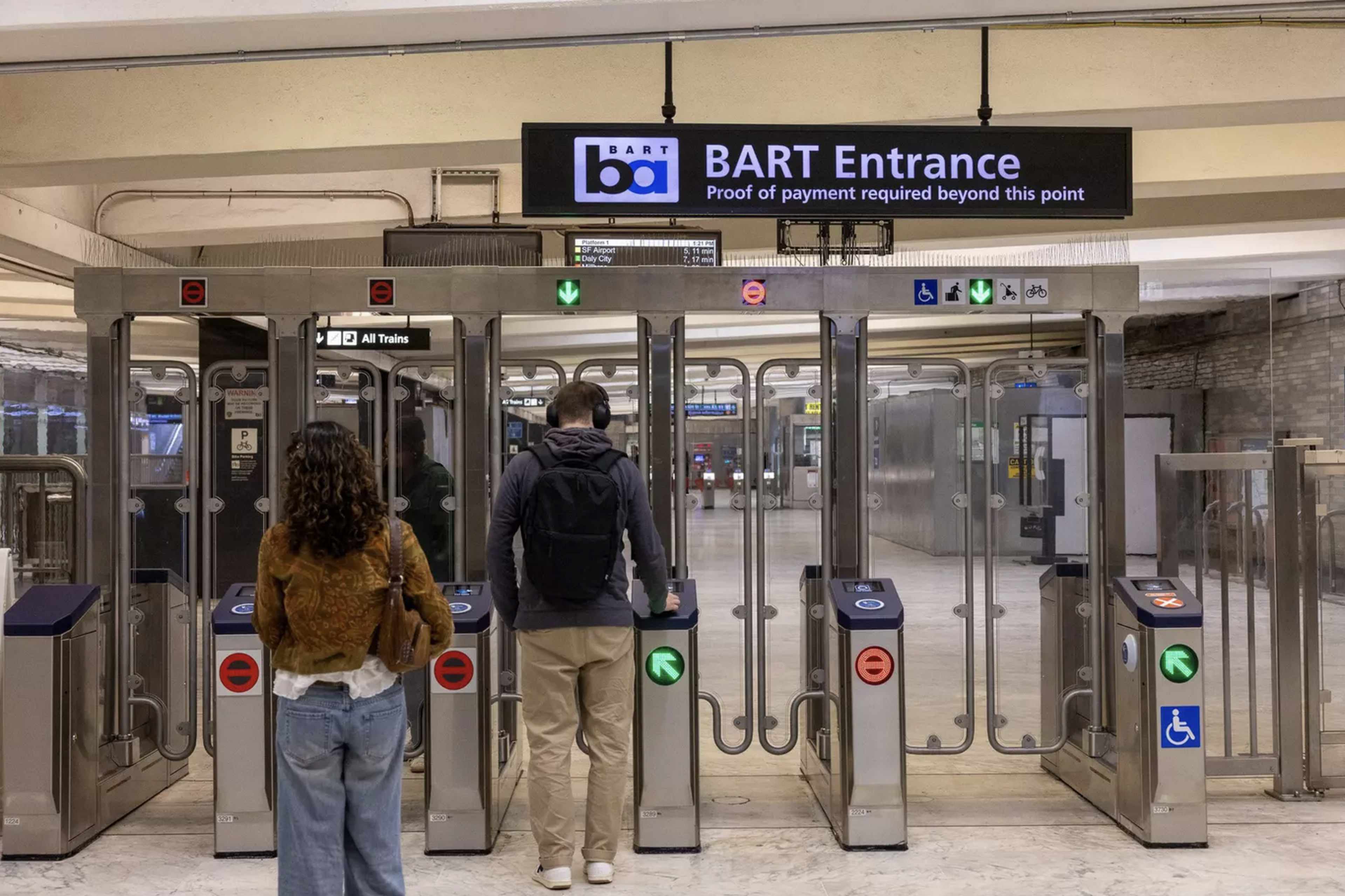 Two people using turnstiles at a BART entrance, with signs indicating payment required and accessible, bike, and priority entry lanes.