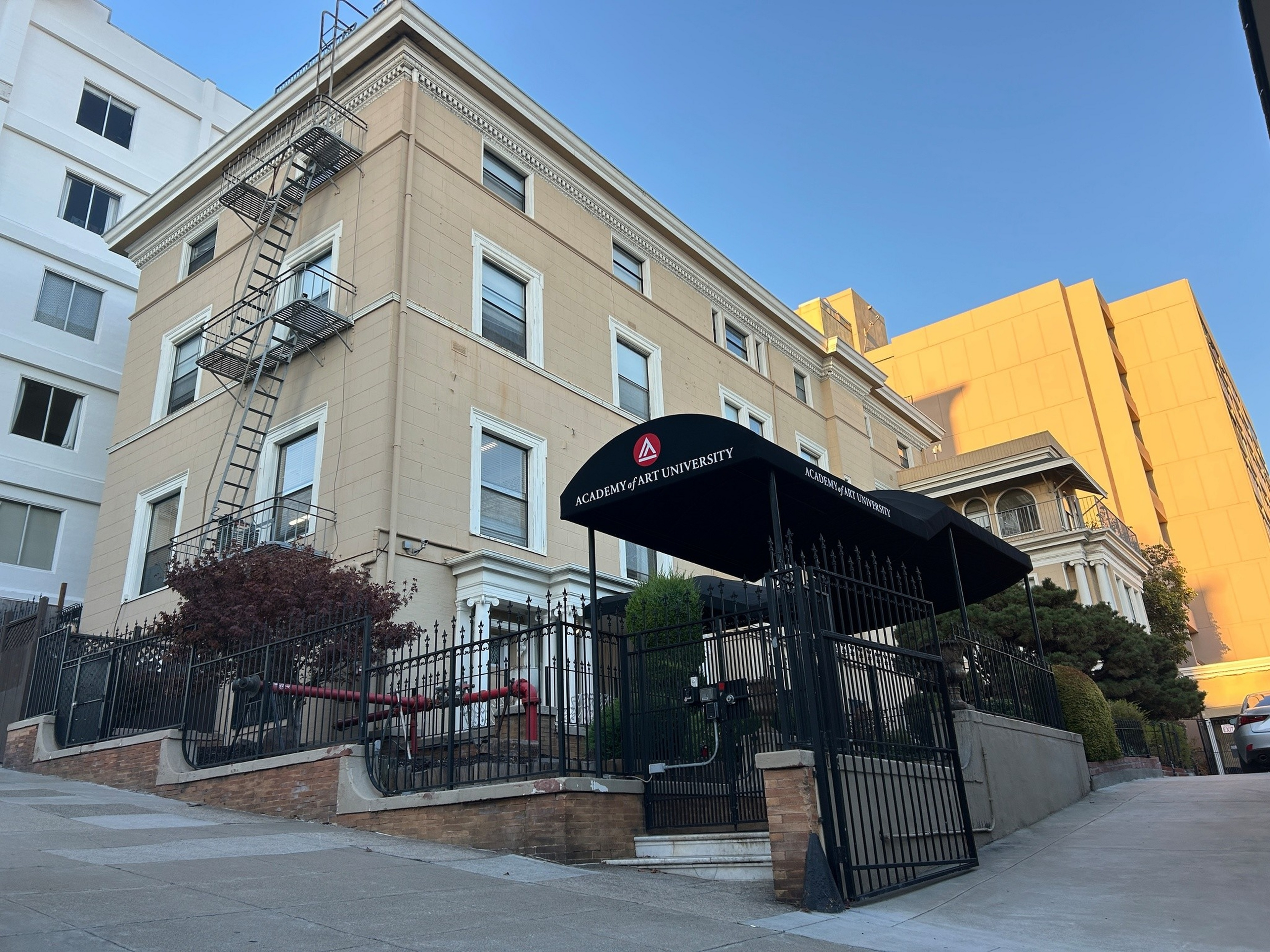 A beige three-story building with a black wrought iron fence and gate, a fire escape, and a black awning labeled "Academy of Art University."