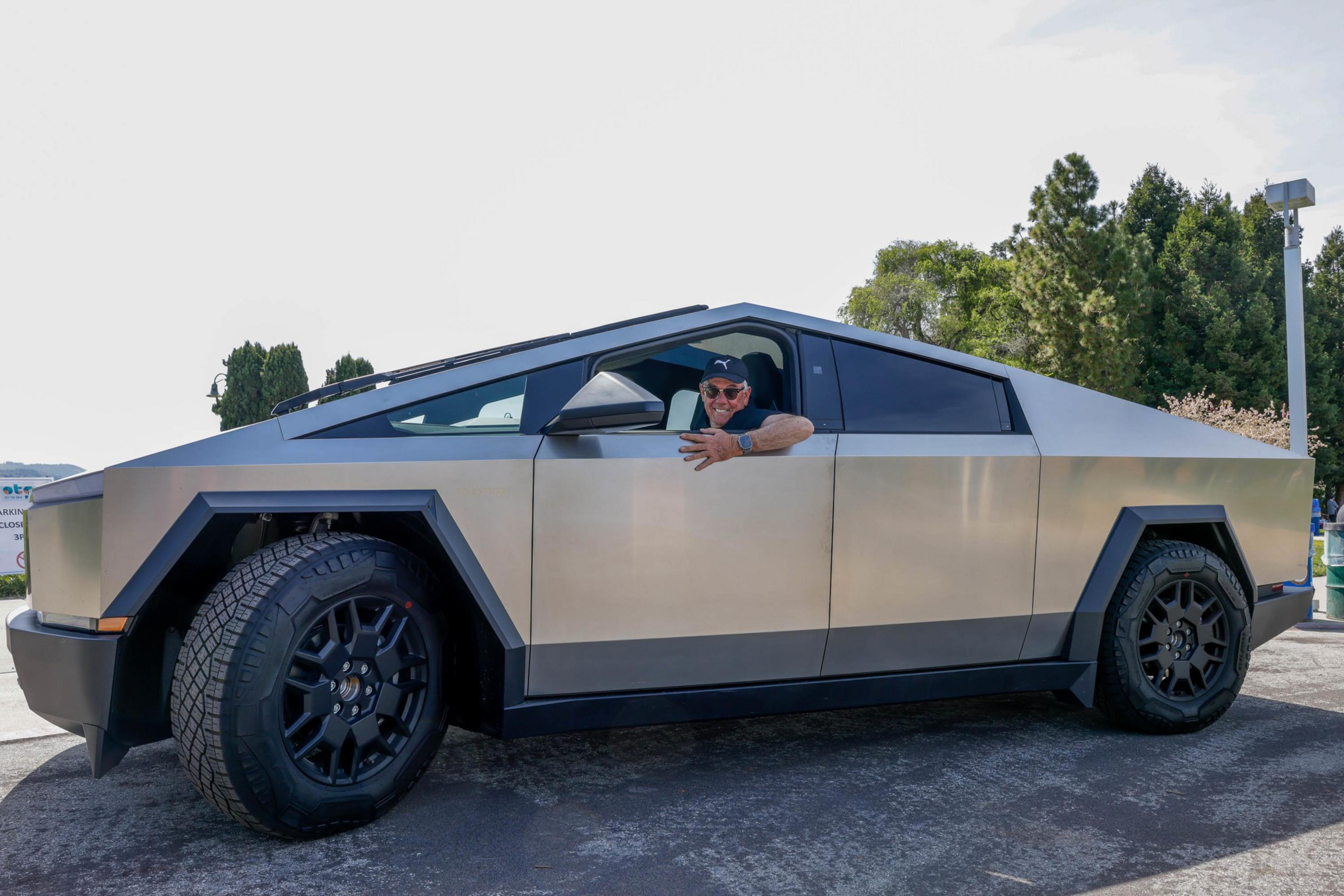 A man in a cap smiles from the driver's seat of a futuristic, angular silver truck.