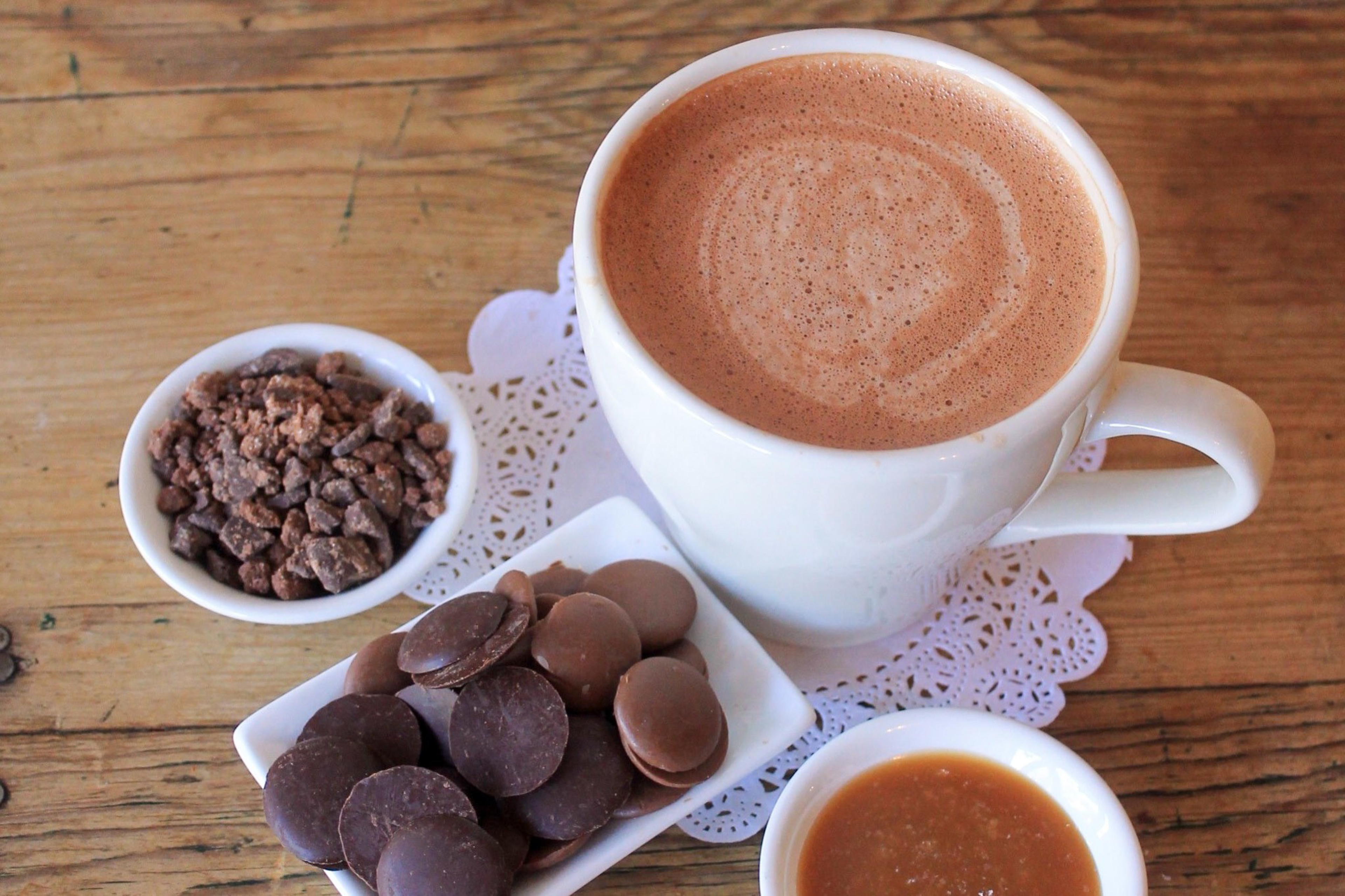 A white cup of frothy hot chocolate sits on a wooden table with small bowls of chocolate chunks, chocolate discs, and caramel sauce nearby.