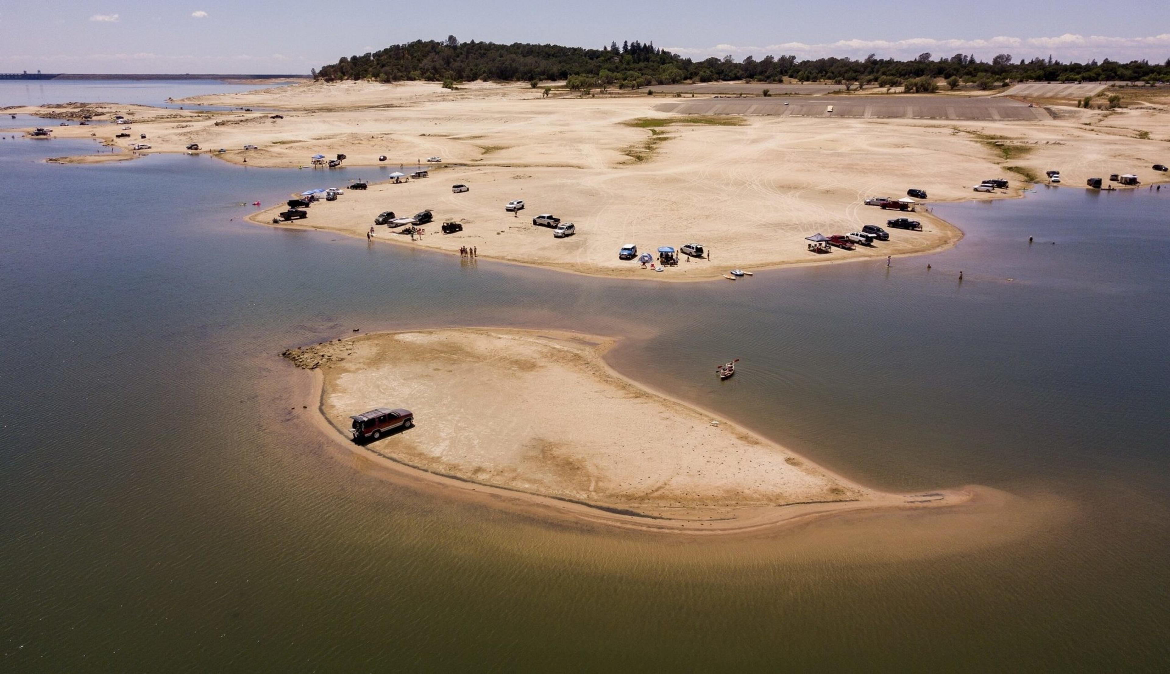 A sandy riverbank with numerous parked vehicles, a small isolated sandy island with one vehicle, and scattered people near the water under a clear sky.