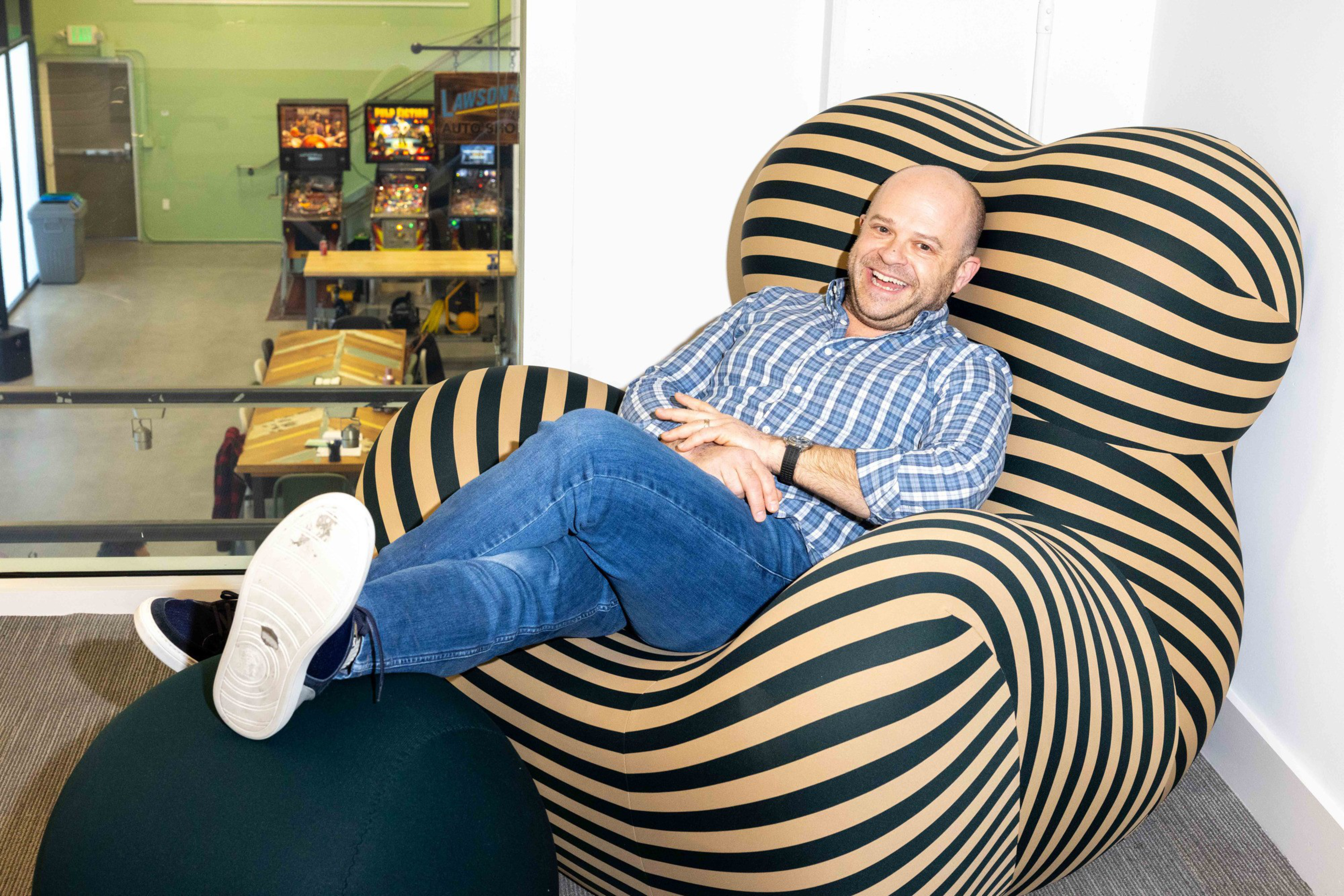 A person in a checkered shirt and jeans is happily lounging on a large, striped chair. In the background, there are pinball machines and a green wall.