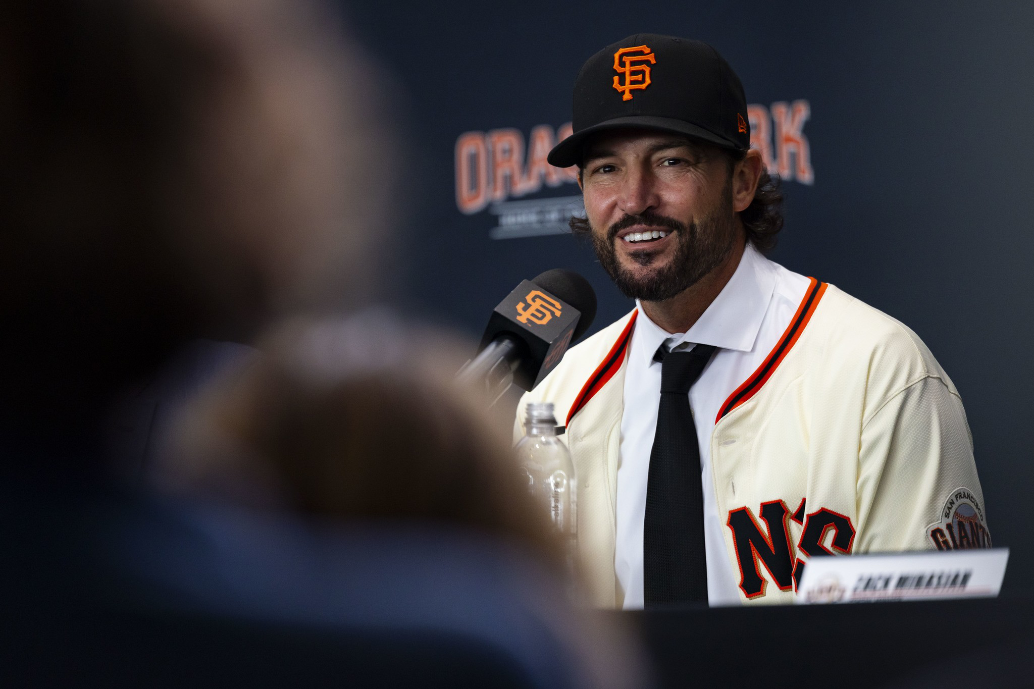 A man wearing a San Francisco Giants baseball cap and jersey smiles while speaking into a branded microphone at a press event.